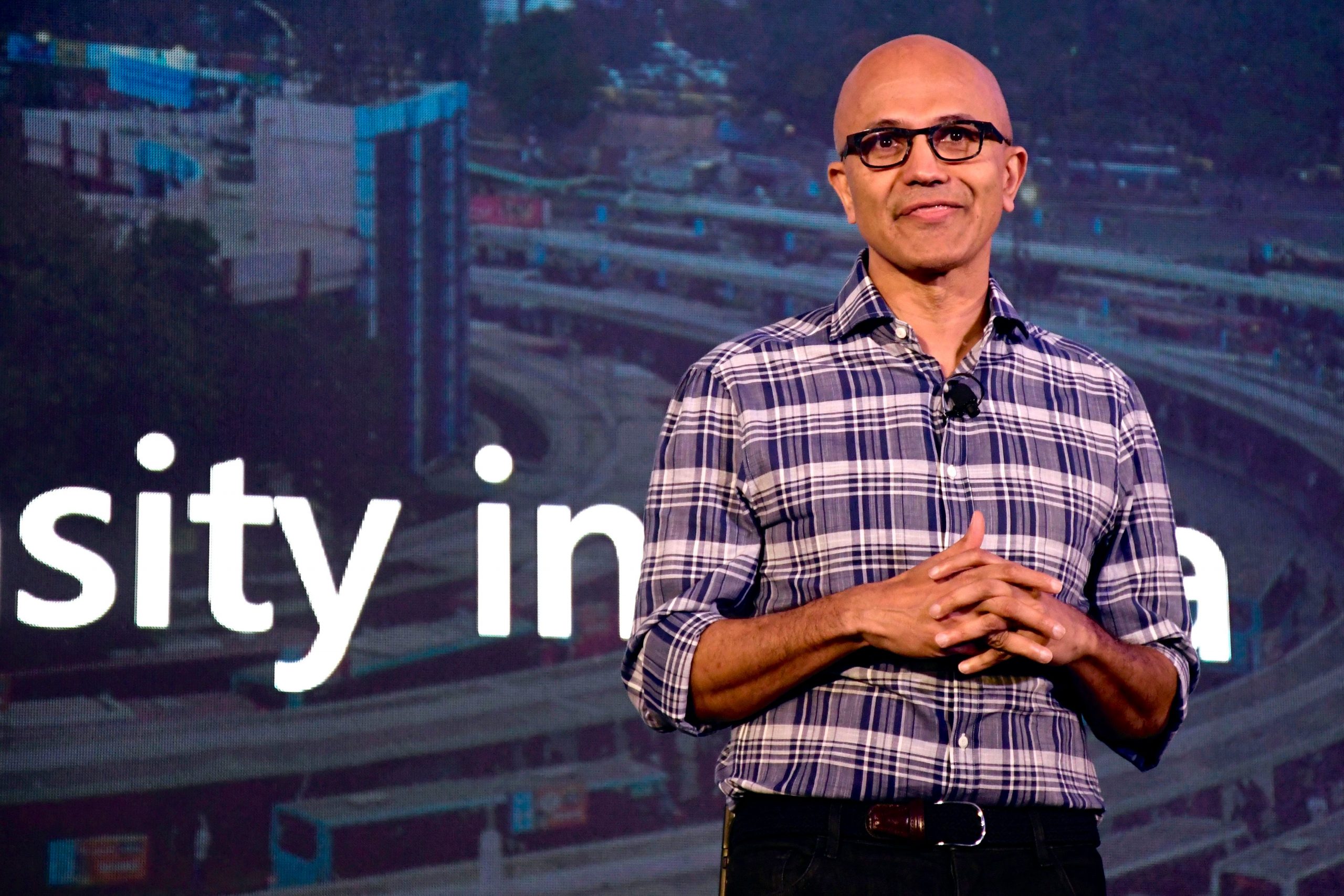 Satya Nadella stand on stage wearing a purple and white checkered shirt.