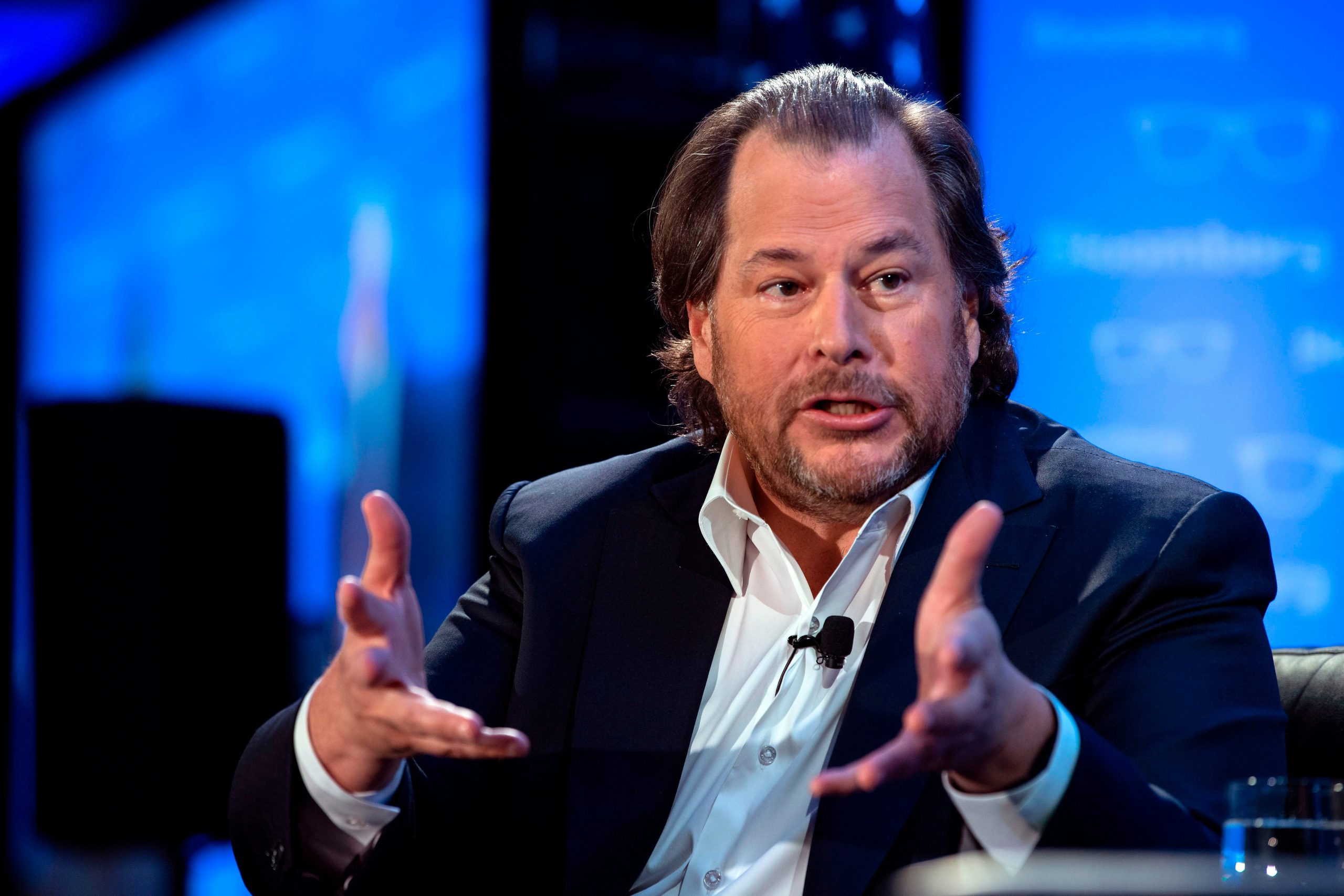 Salesforce founder Marc Benioff, wearing a sports coat jacket and in front of a blue background on a stage, extends his hands forward during a talk.