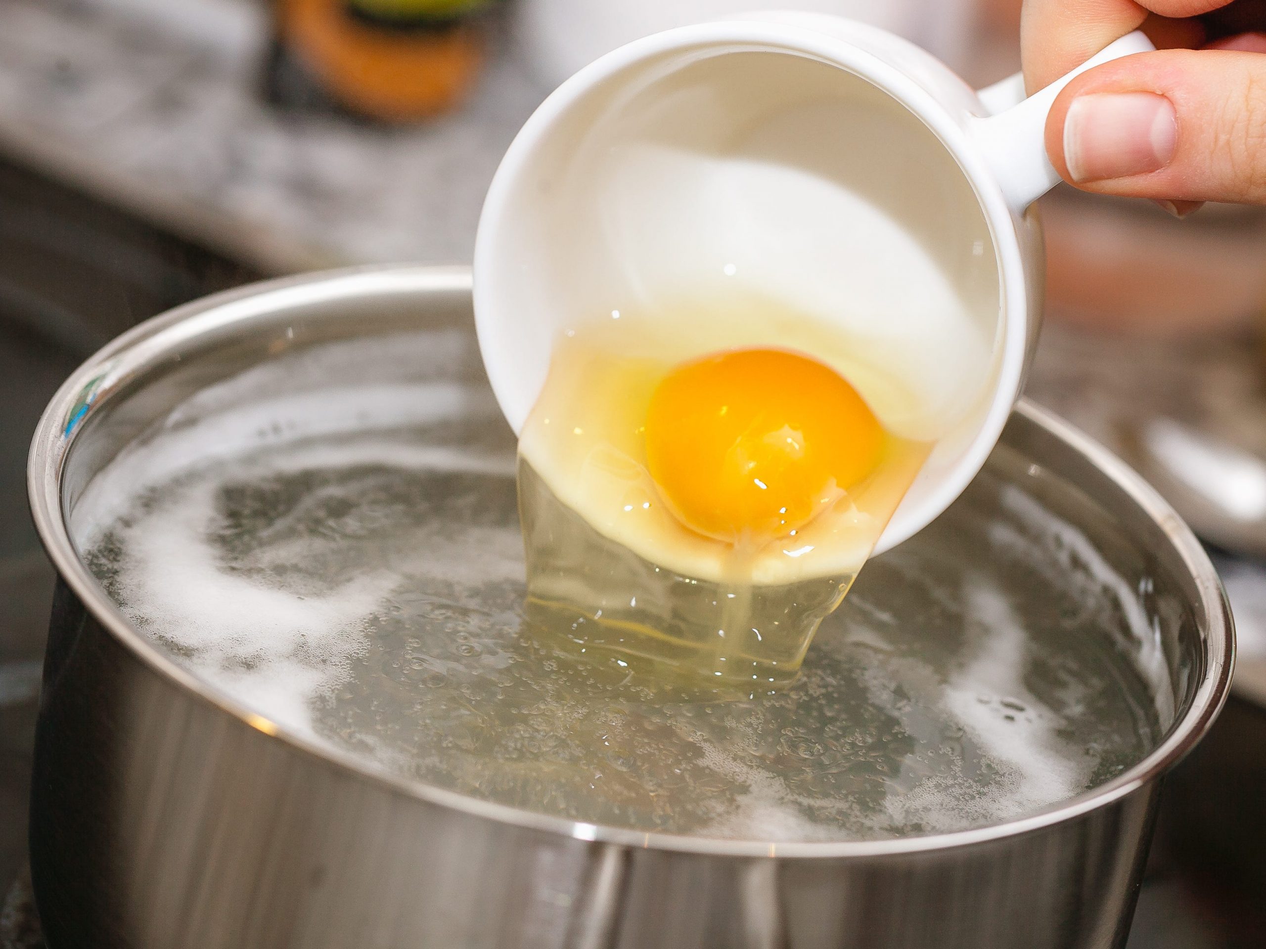 A hand pouring an egg out of a teacup into a pot of boiling water