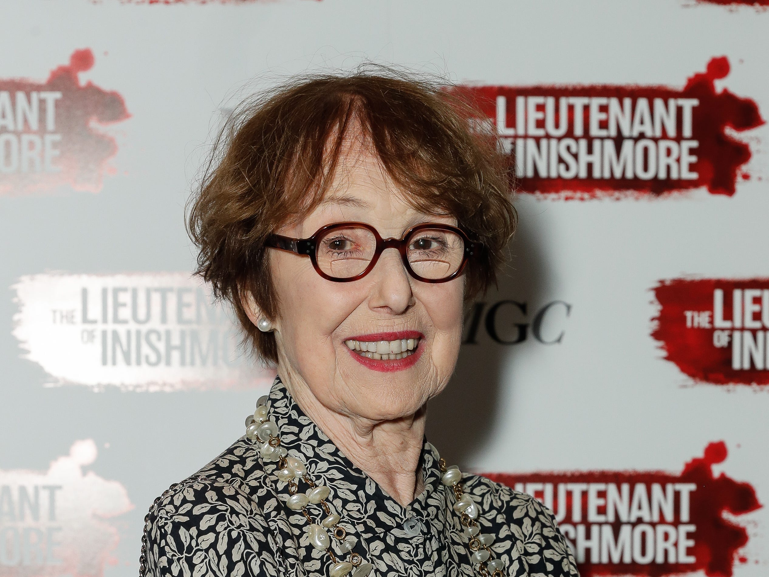 Una Stubbs smiles in glasses in front of signs for The Lieutenant of Inishmore