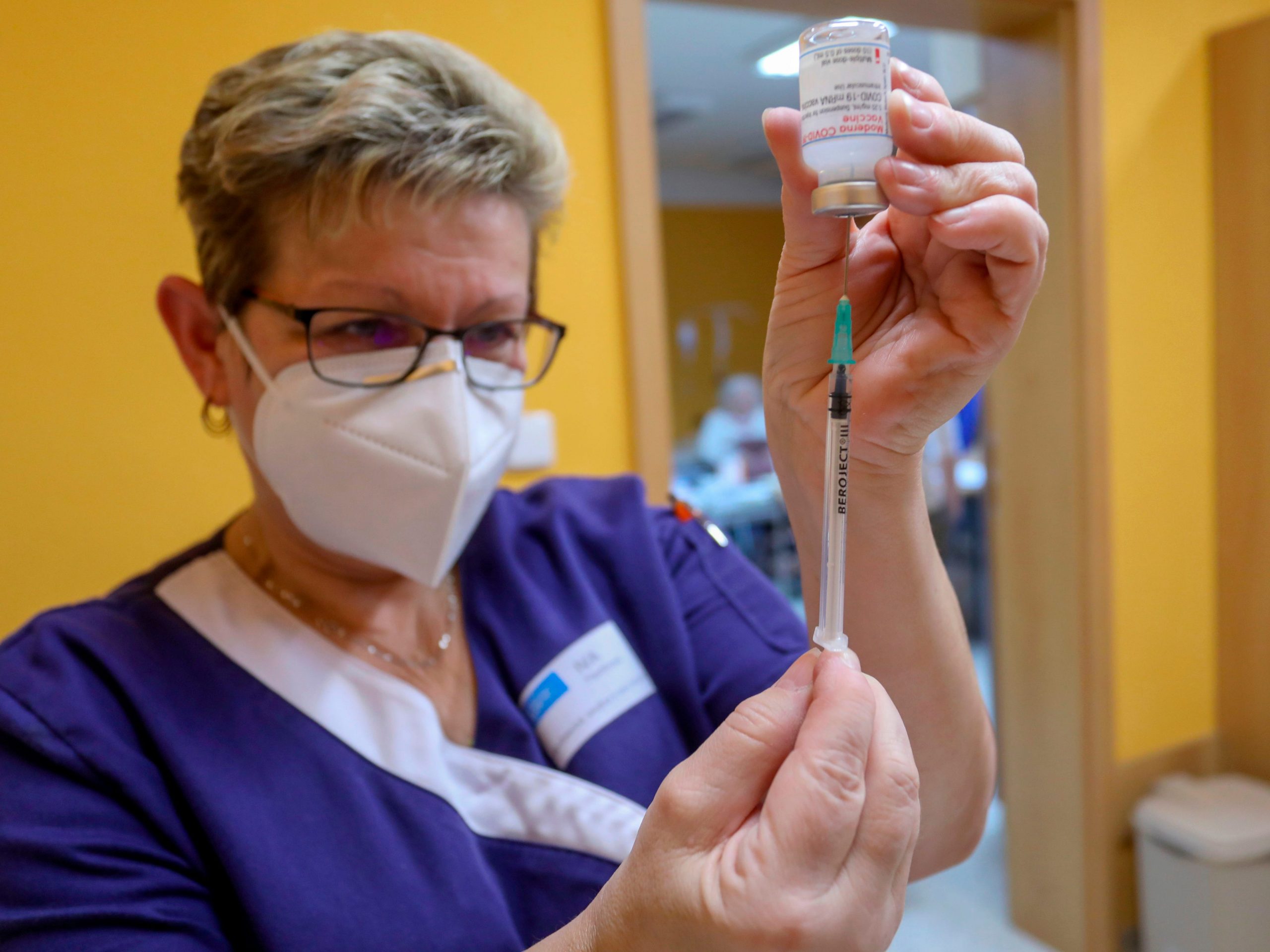A healthcare professional wearing a mask fills a syringe of the Moderna COVID-19 vaccine from a glass vial.