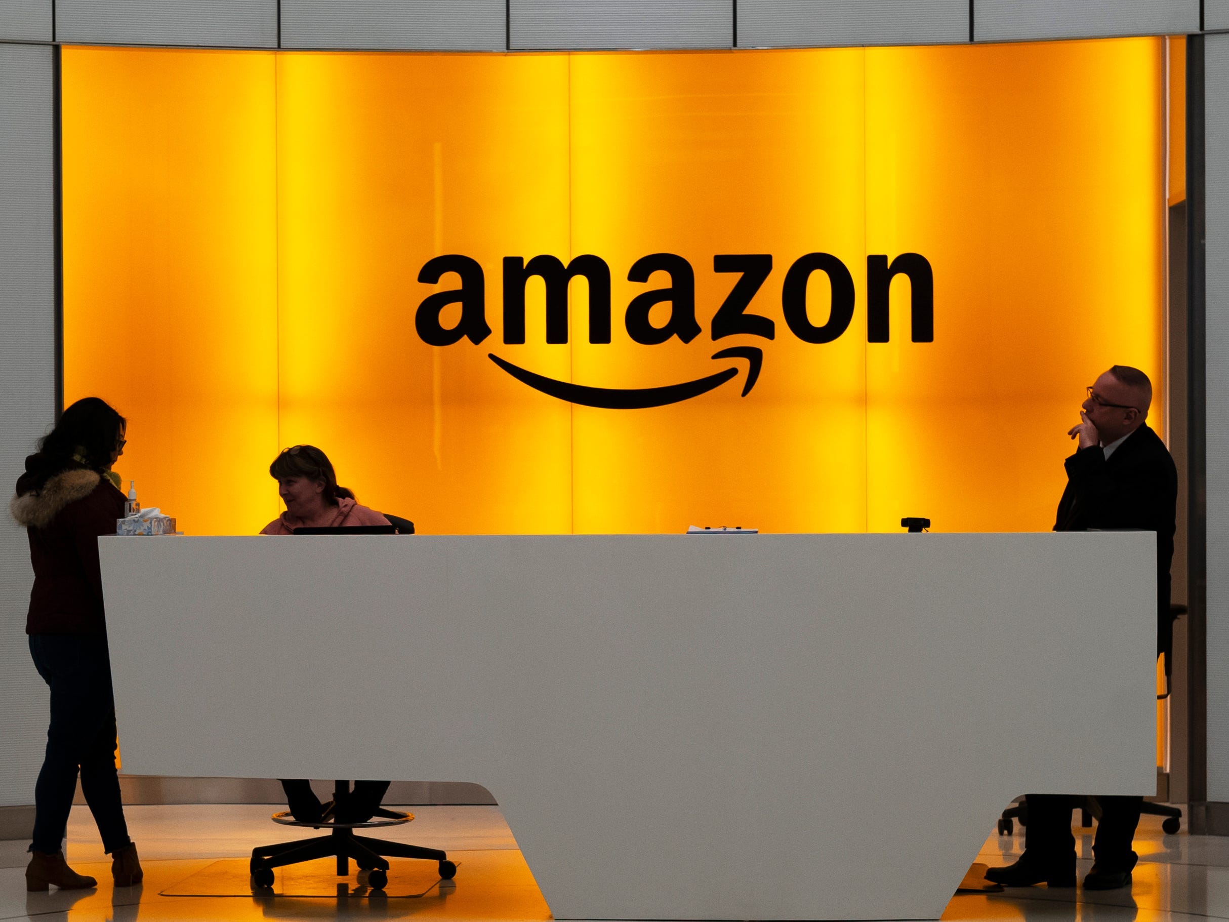 Amazon office front-desk staff stand in front of an orange sign with the Amazon smile logo