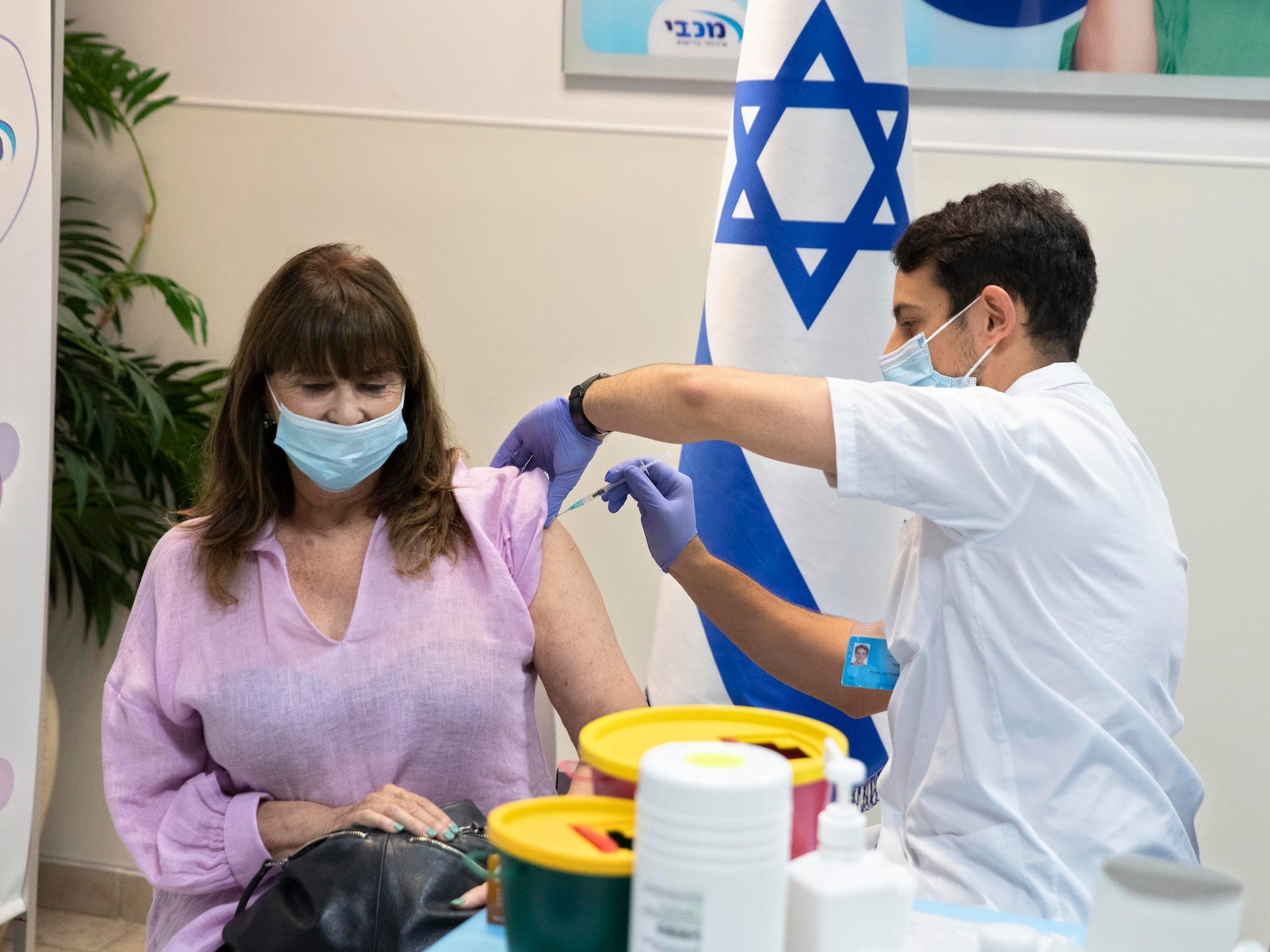 A masked woman wearing a purple shirt is getting vaccinated by a man in scrubs wearing a mask in front of an Israeli flag