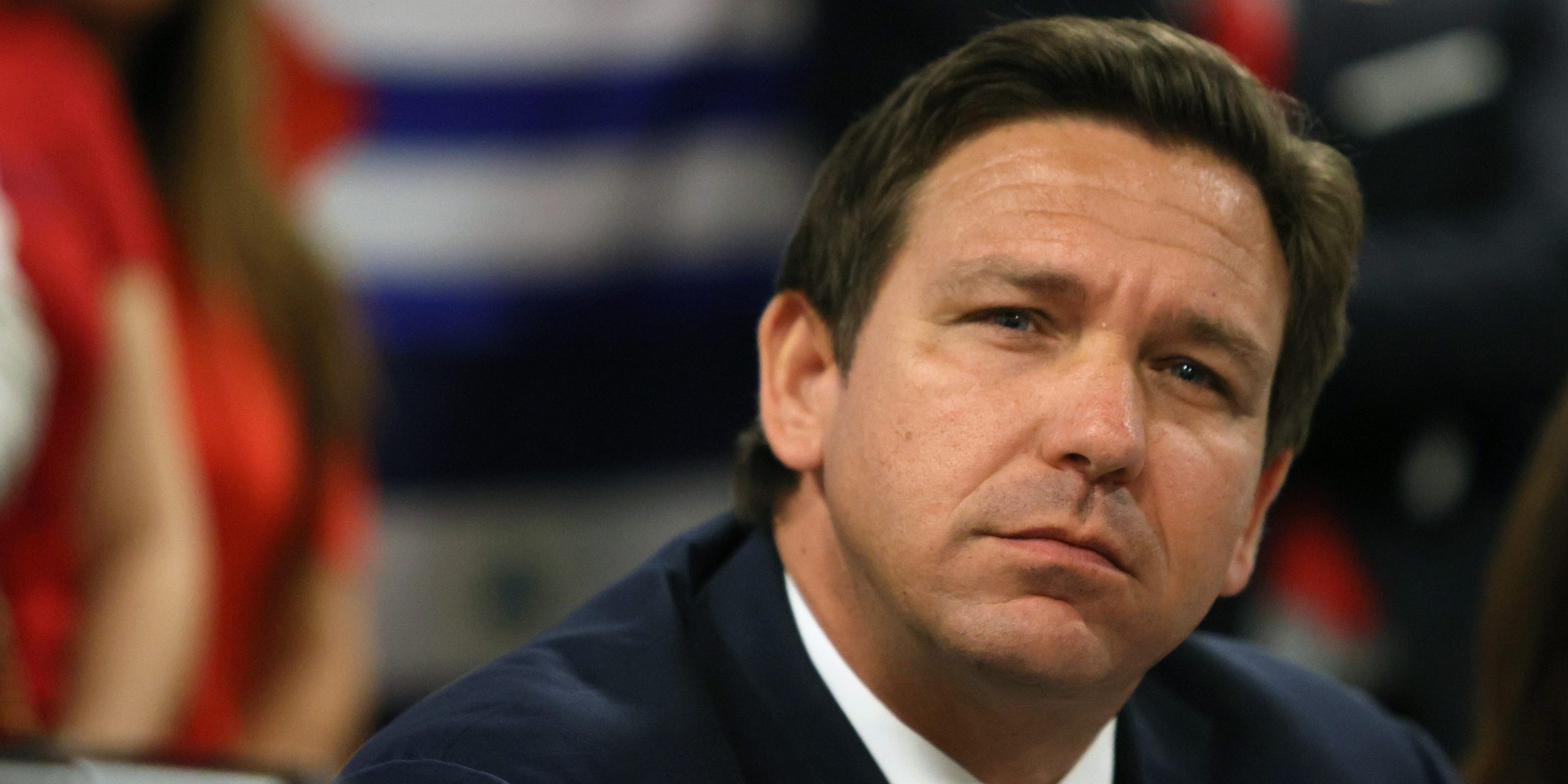 Florida Governor Ron DeSantis wears a black suit and blue tie and looks into the distance.