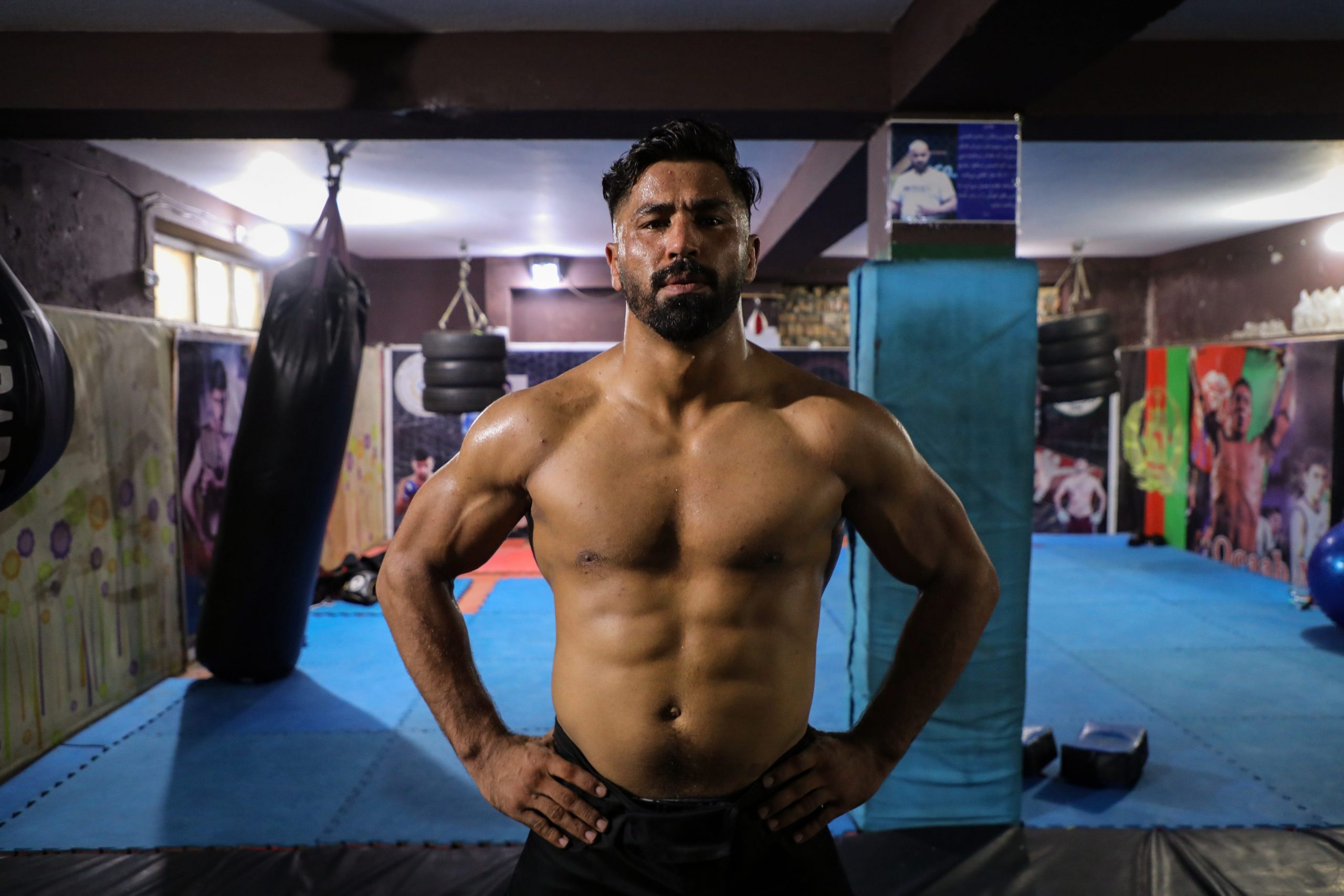 A man stands in a spare, dimly-lit gym, shirtless and with his hands on his hips.