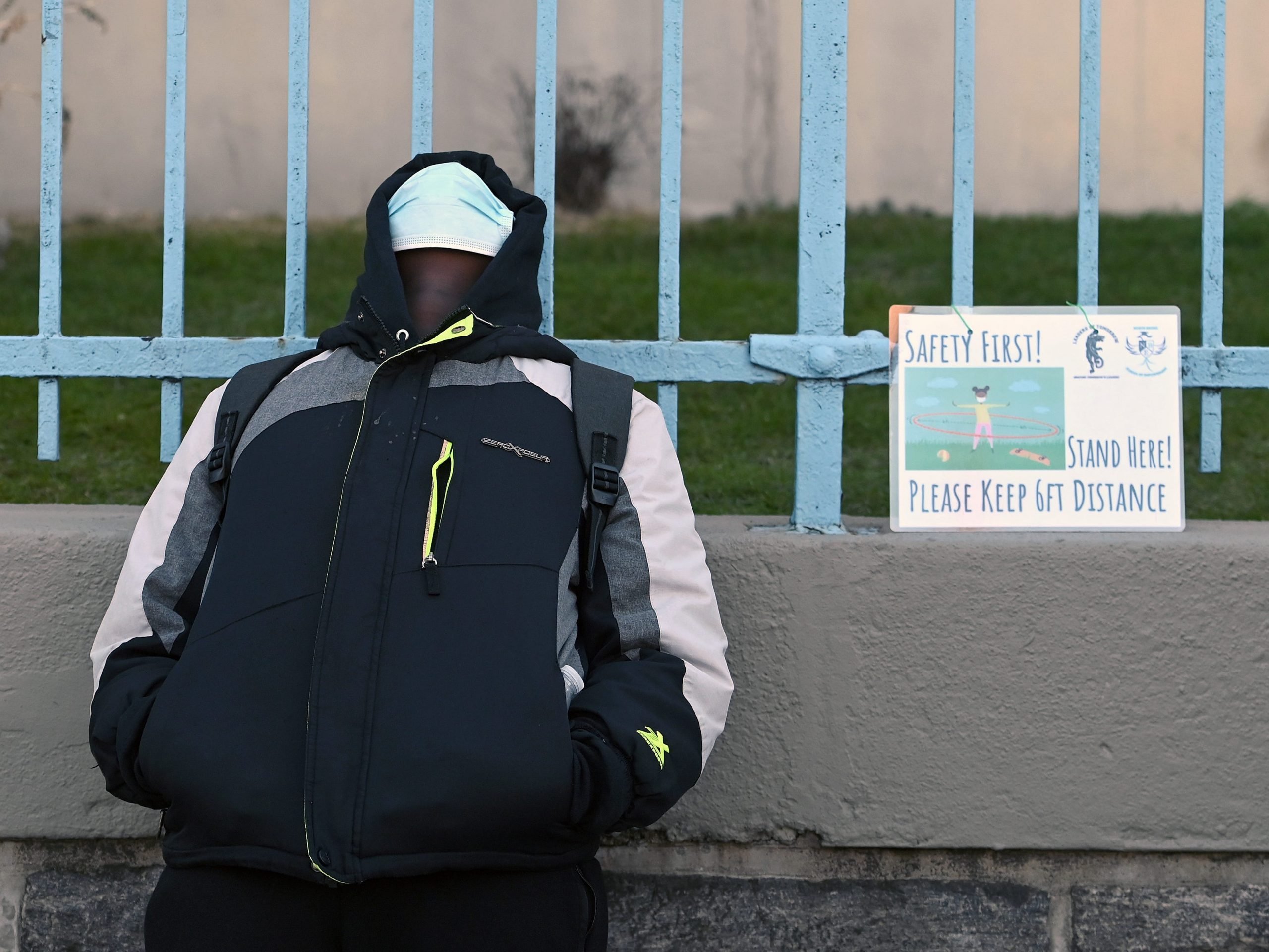 a student stands against school walls next to a sign that reads 'safety first! keep social distance'