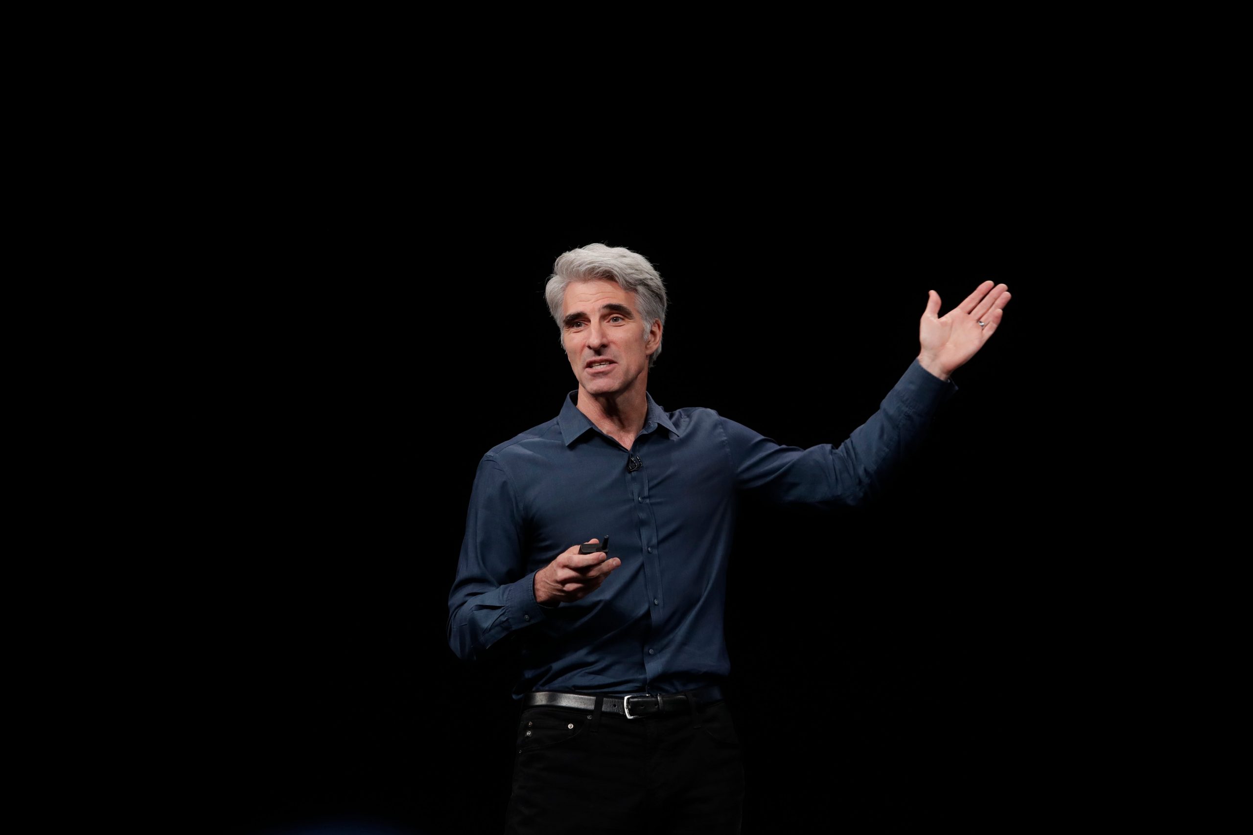 Apple SVP Craig Federighi raises his hand as he speaks on stage in front of a black background