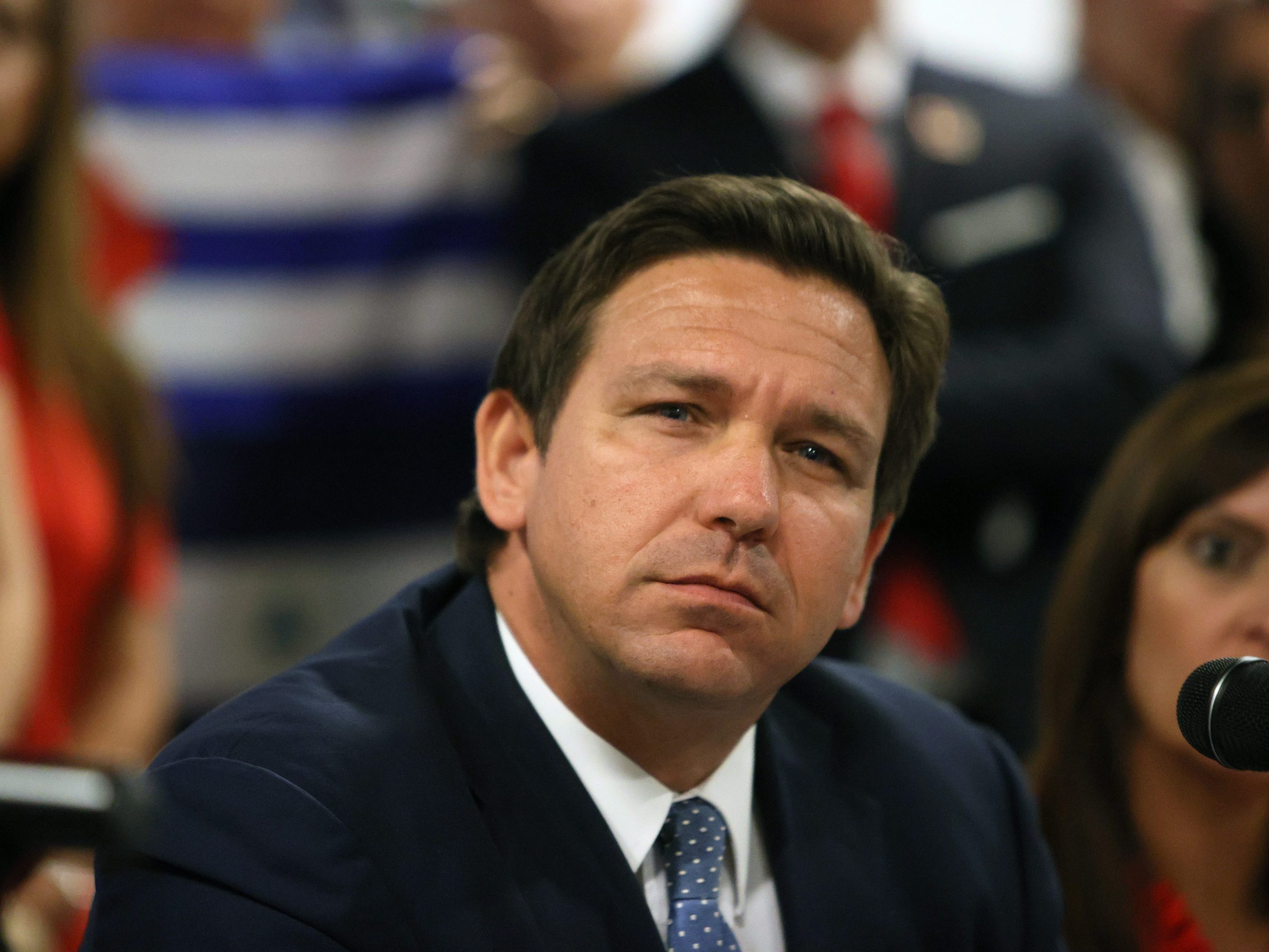 Florida Governor Ron DeSantis wears a black suit and blue tie and looks into the distance.