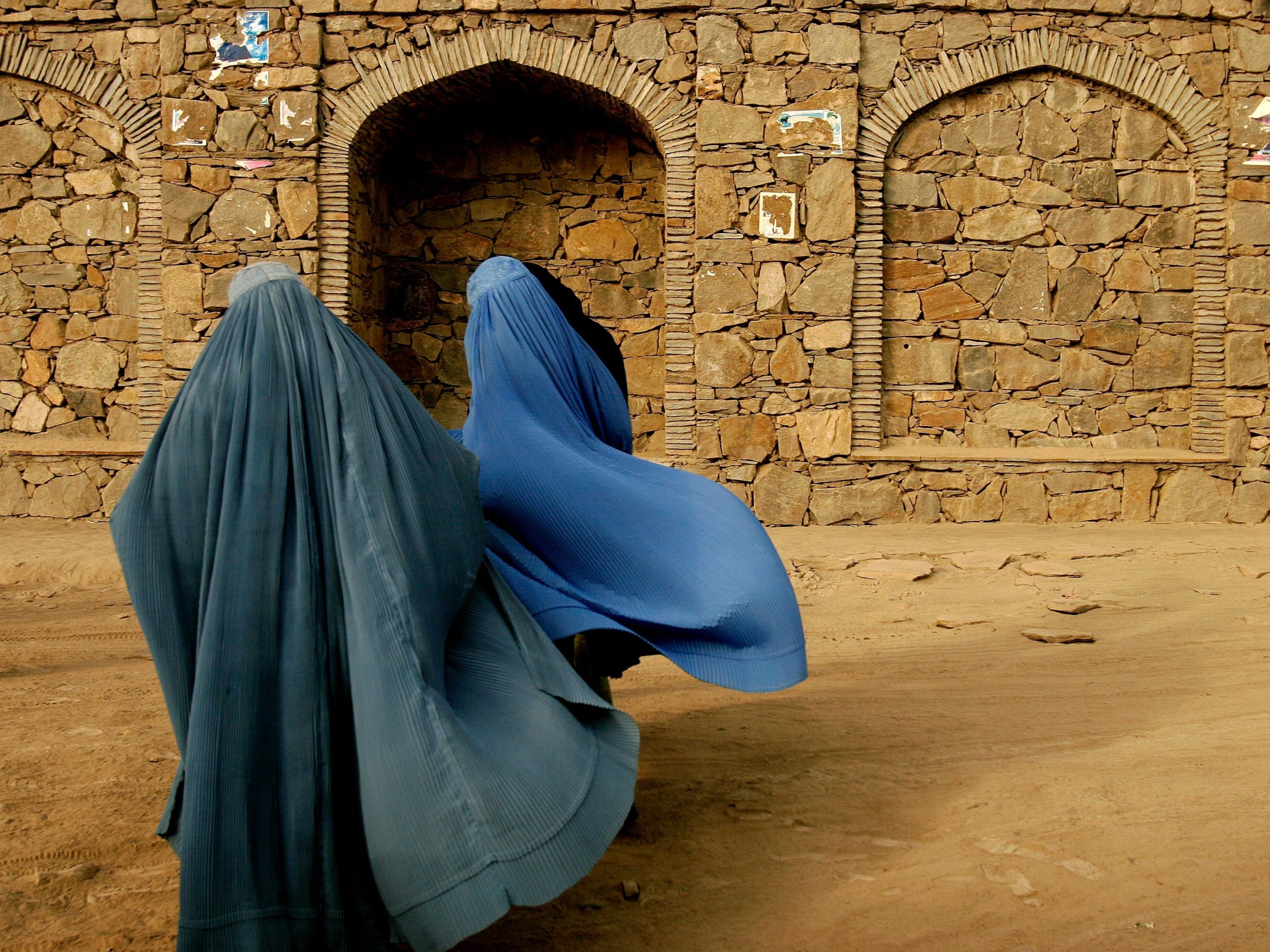 Two women wear blue burkas covering their entire bodies in kabul with a stone wall behind them