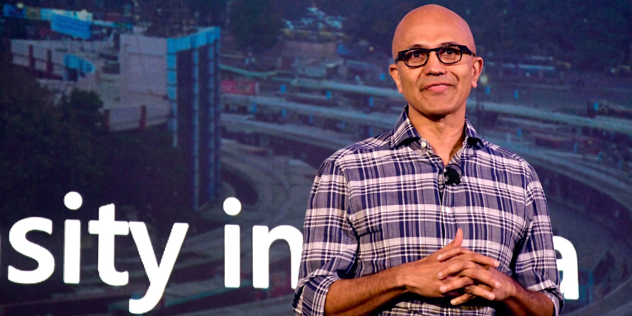 Satya Nadella stand on stage wearing a purple and white checkered shirt.