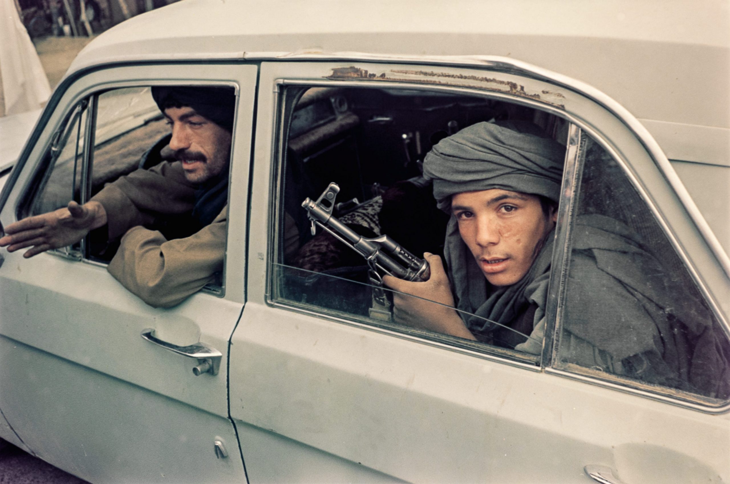 Two men are seen in a car, and the one in back looks out the window as he clasps a gun.