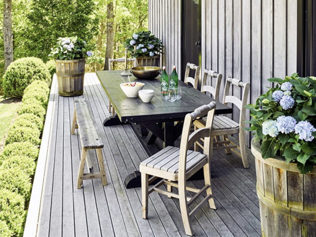 A large wooden dining table surrounded by chairs on a wooden front porch