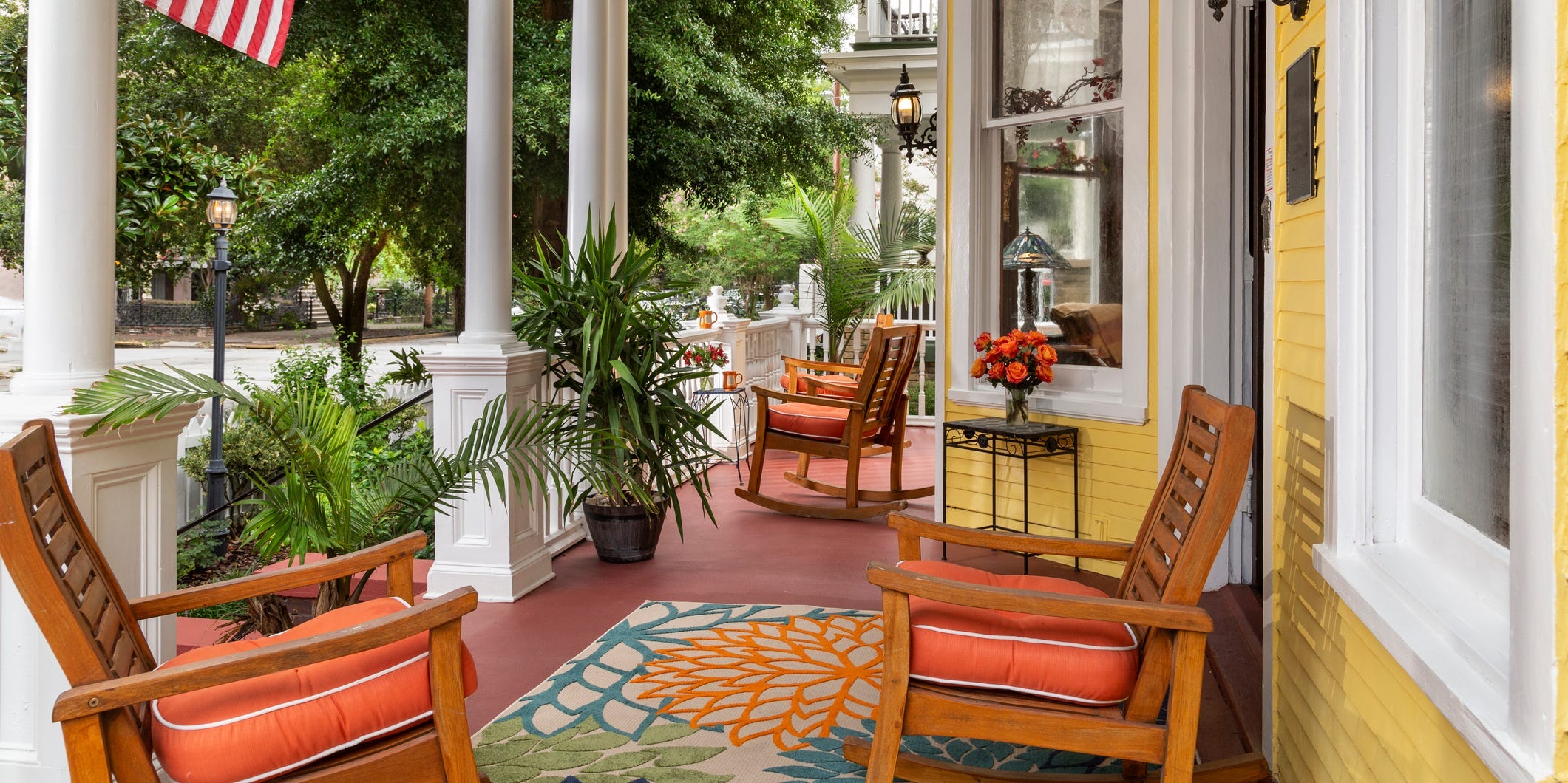 A colorful front porch decorates iwth several wooden chairs with orange cushions, a blue rug, and palm plants