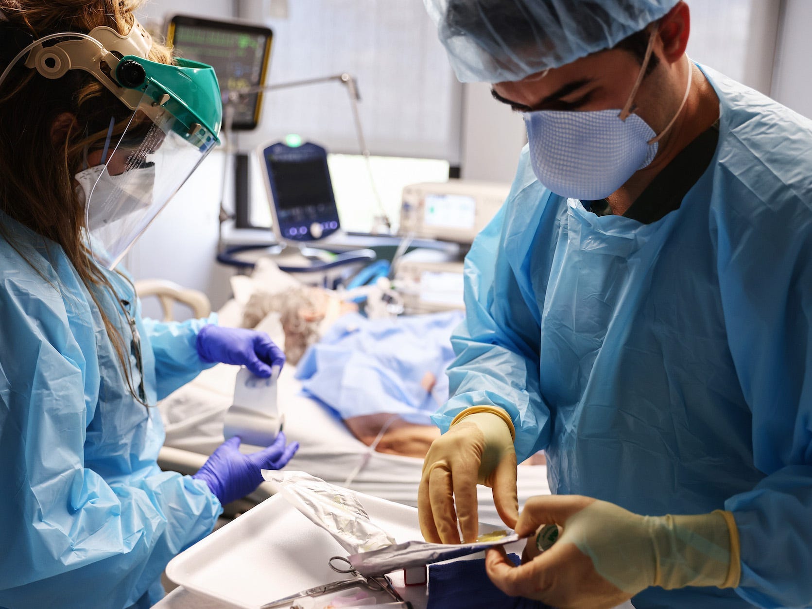 A clinician is looking over his tools while a nursing writes something down. Both are in full PPE. In the background is a COVID-19 patient.