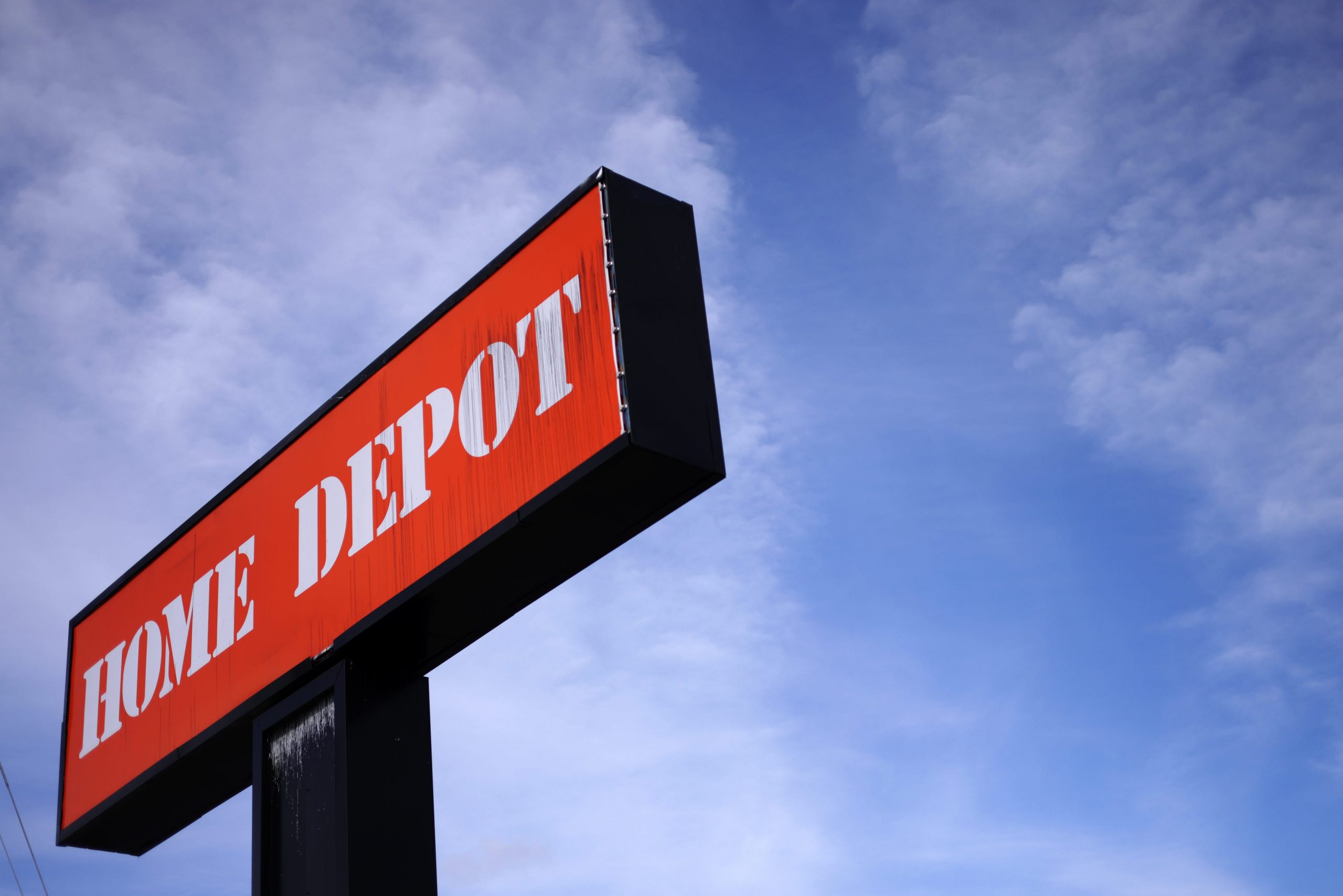A bright orange sign for Home Depot stands tall against a blue sky with clouds.