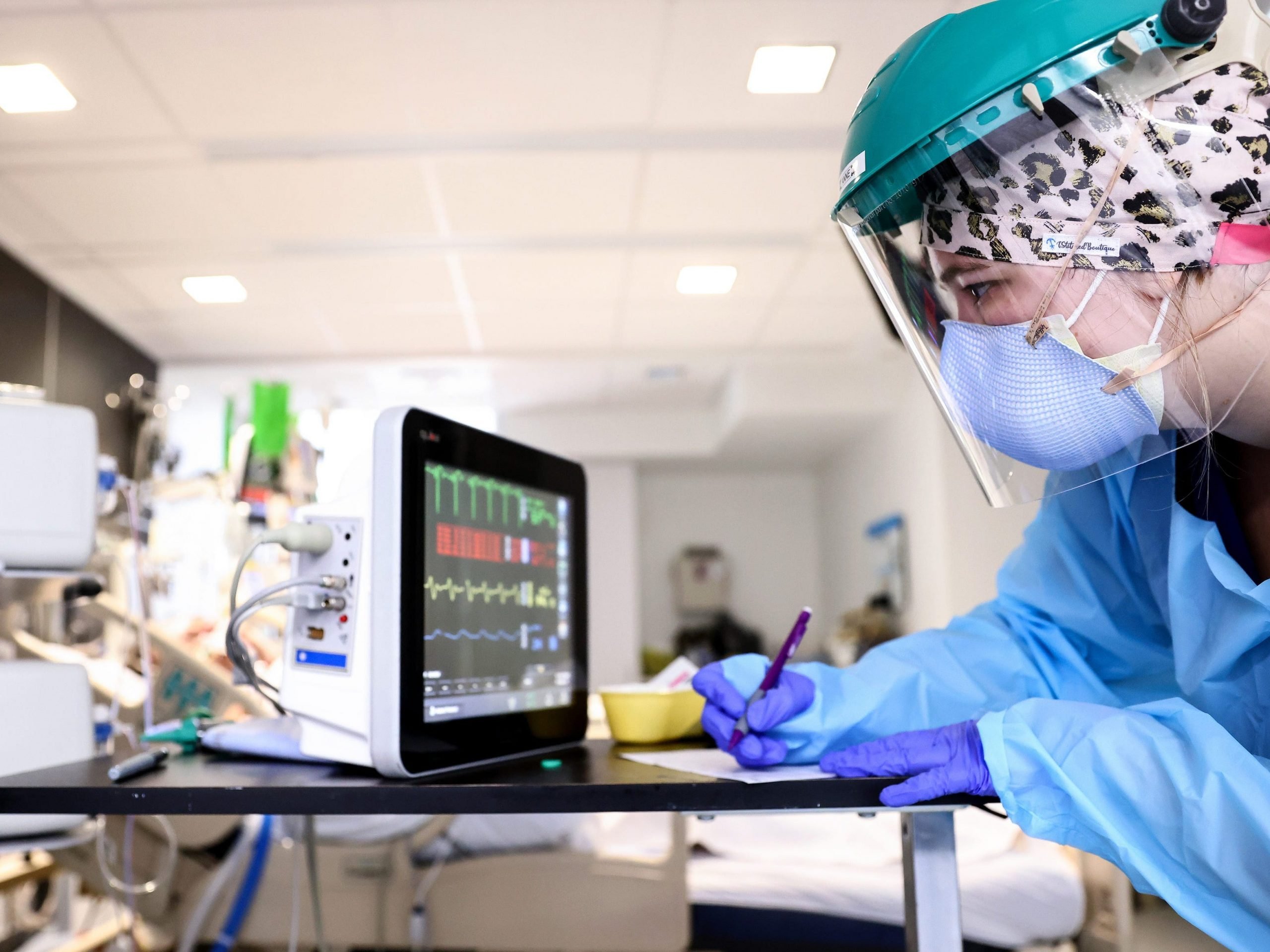 A woman wearing a mask and face shield and PPE takes readings from a hospital instrument