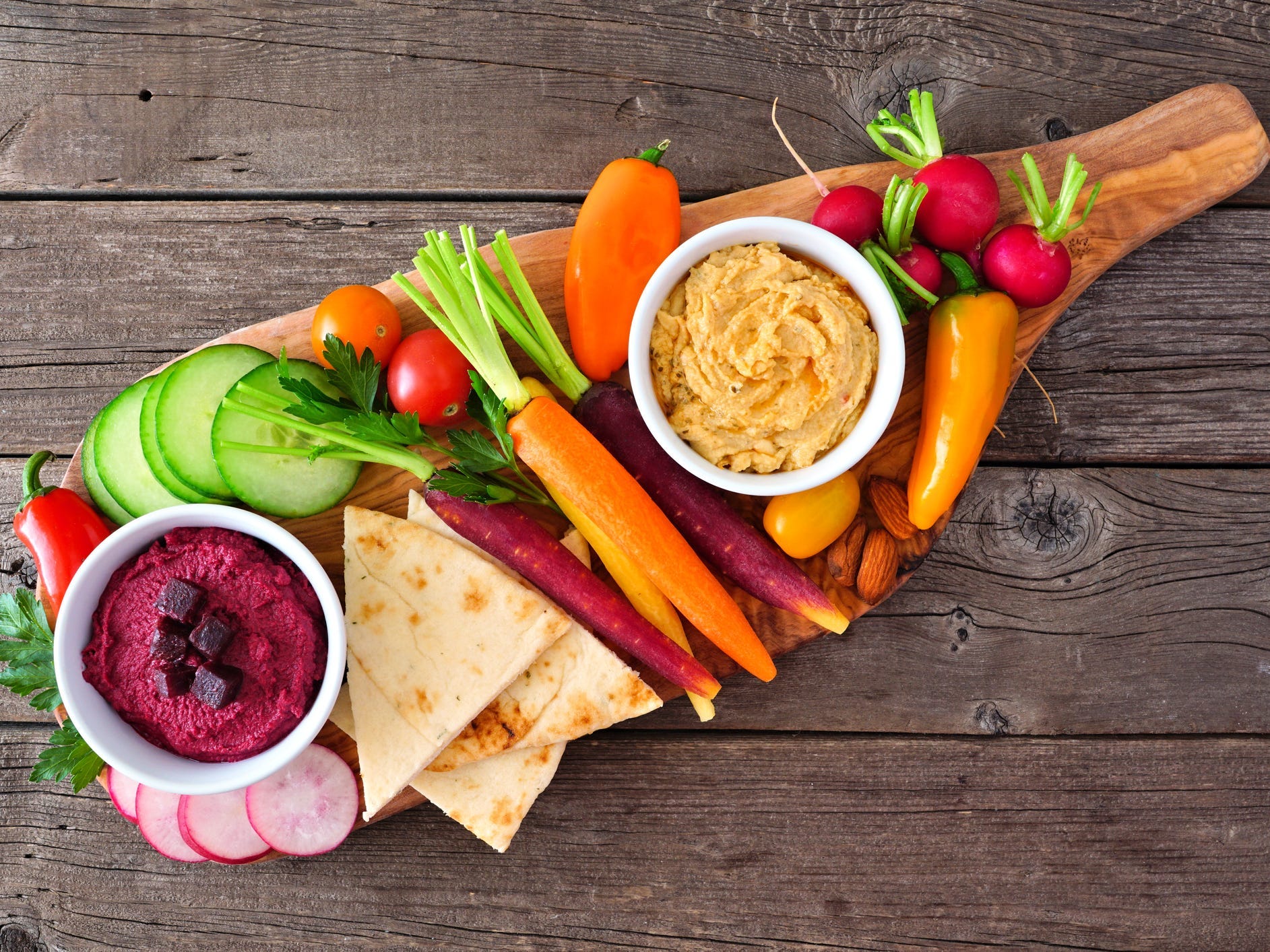 A board with hummus, cucumbers, peppers, carrots, pita bread, and other items.