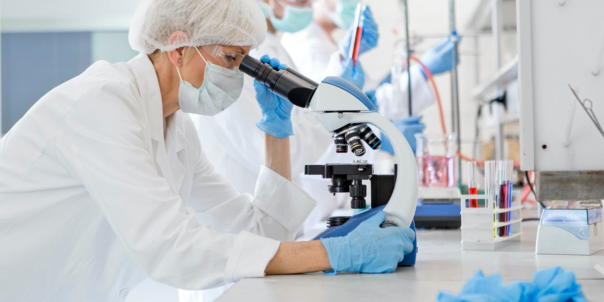 A scientist, who is wearing blue gloves and a lab coat, looks through a microscope. There are other scientists working next to her.