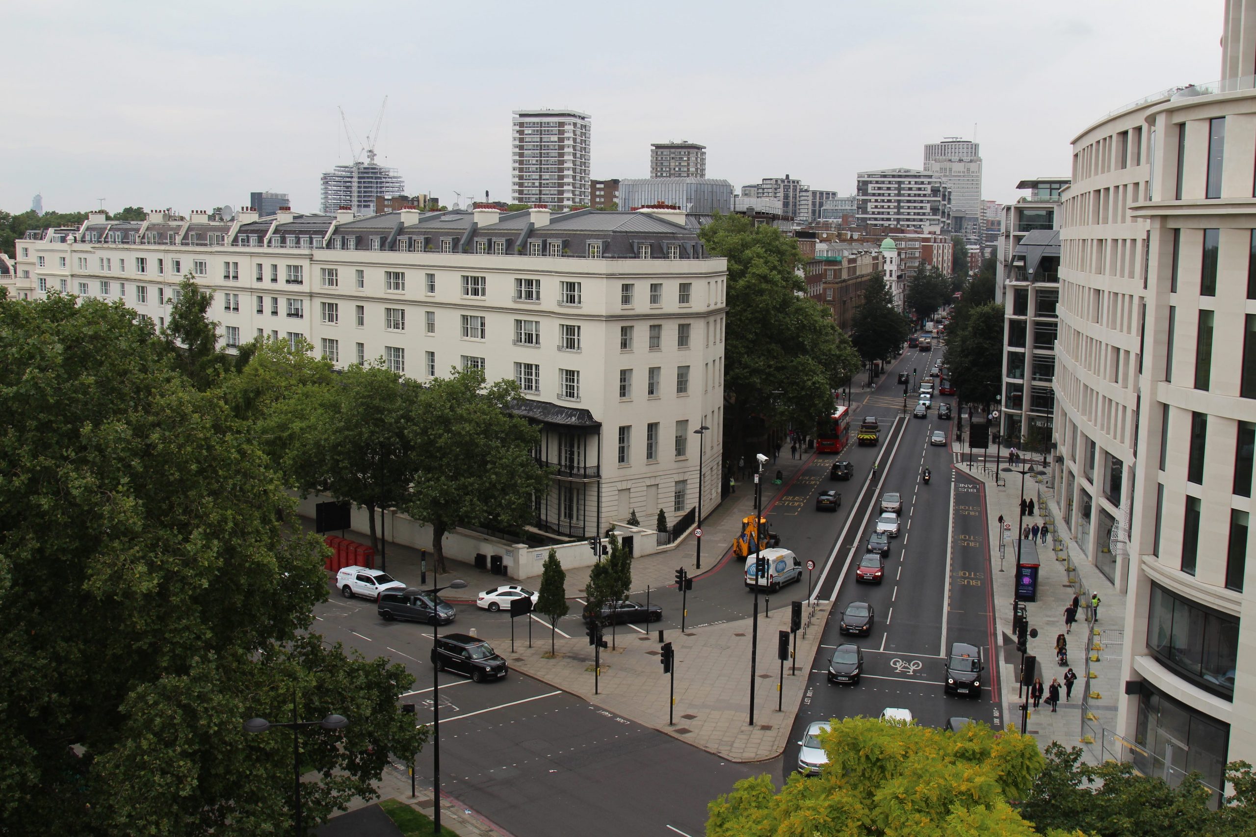 A collection of white buildings in London's busy city center with thick green trees lining the sides of the roads.