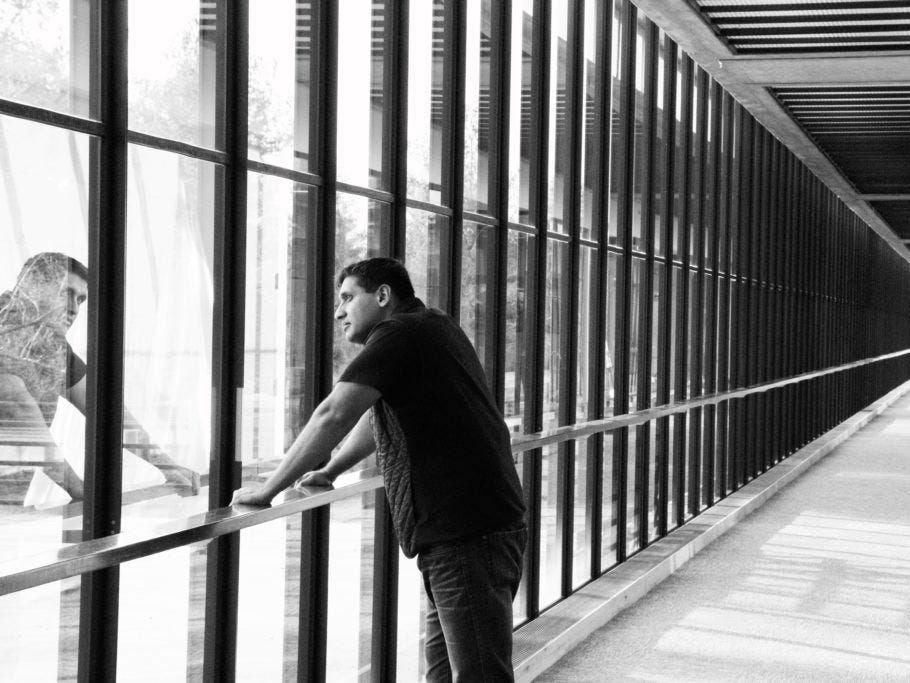 A man wearing a black t-shirt leans on a railing and looks out of a window.