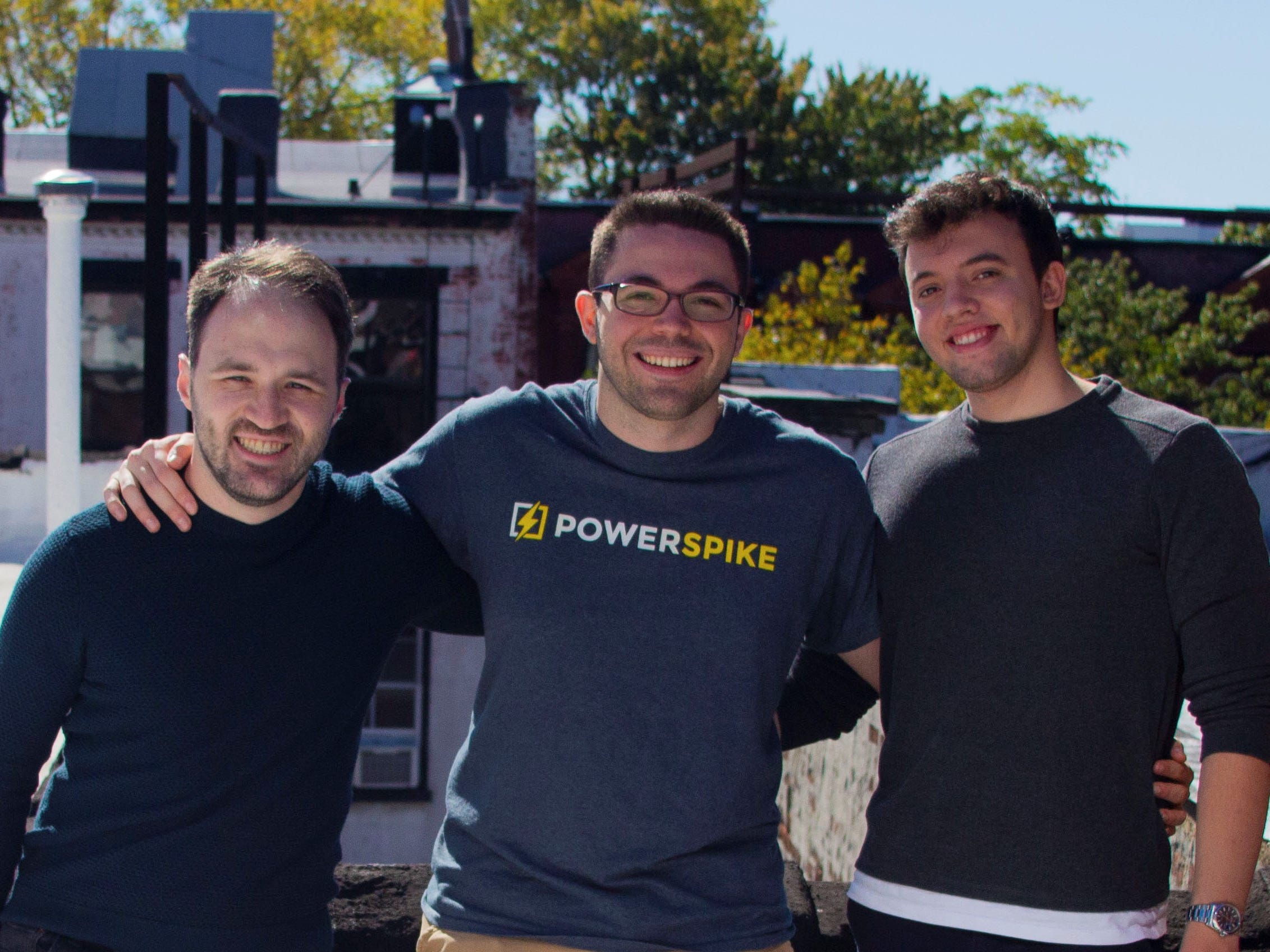 AJ Damiano, Eric Rice and Michael Paris standing outside, smiling with arms around each other's shoulders. AJ is wearing a blue shirt with the Powerspike name and logo on it