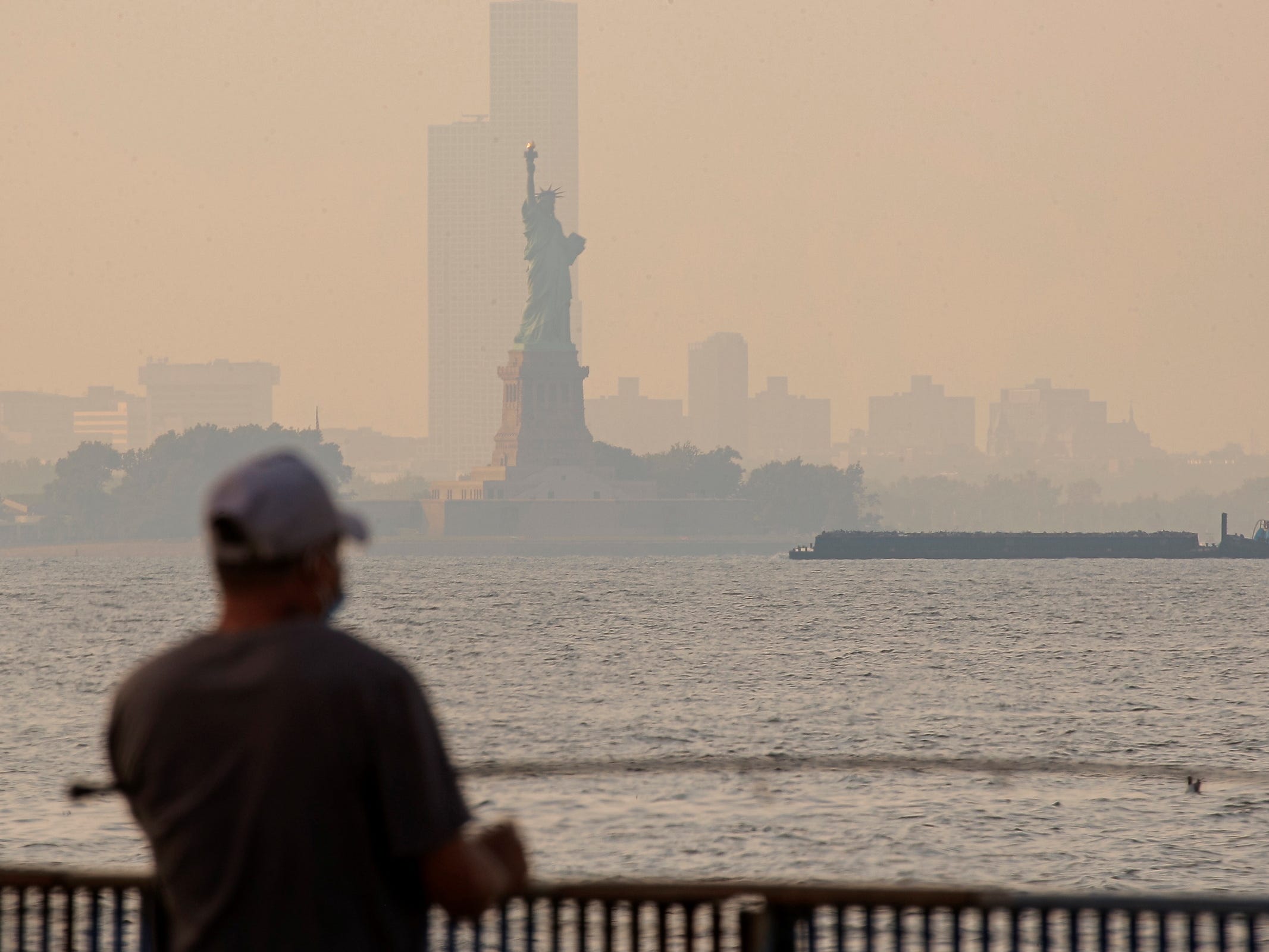 man in baseball cap looks across harbor at statue of liberty through orange haze of wildfire smoke