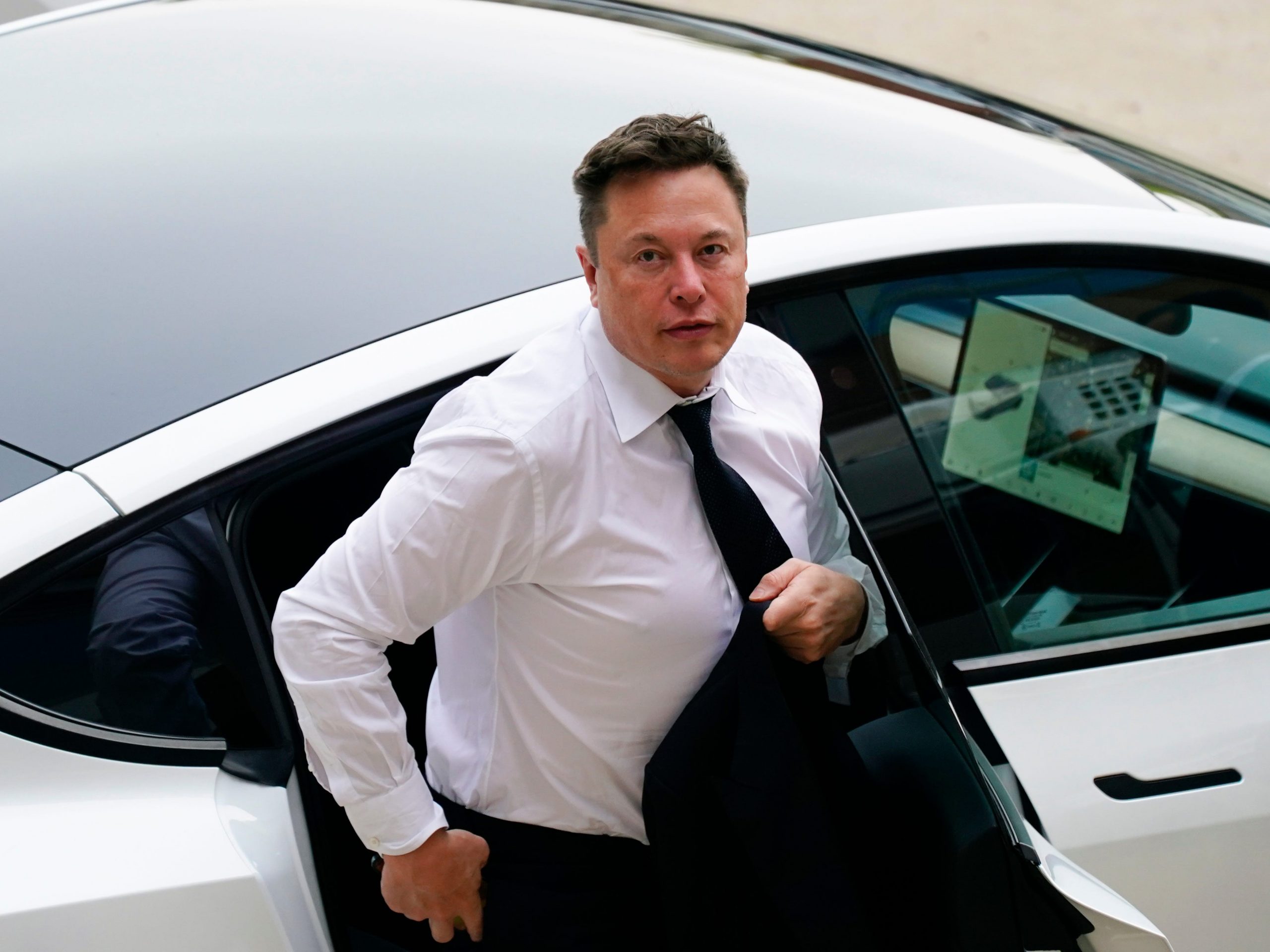 Tesla CEO Elon Musk stepping out of a silver Tesla wearing a white shirt and black tie on a sunny day
