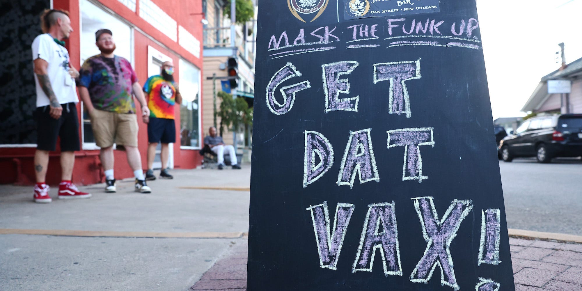 A chalboard sign has an inscription scribbled on it in chalk. People are waiting in the background in front of a red building