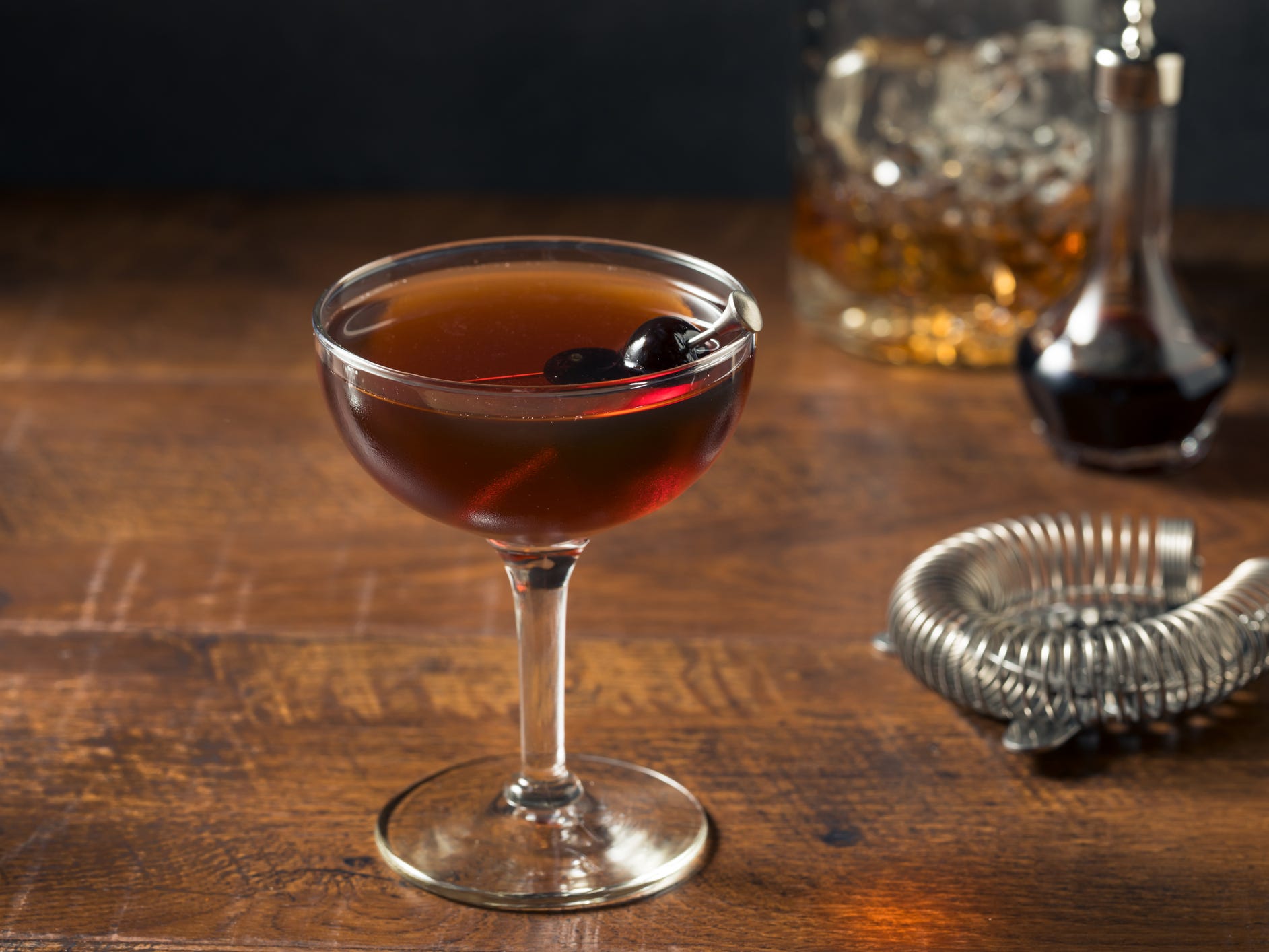 A Manhattan cocktail in a coupe glass garnished with a cherry. A cocktail strainer and a glass decanter of whiskey sit in the background.