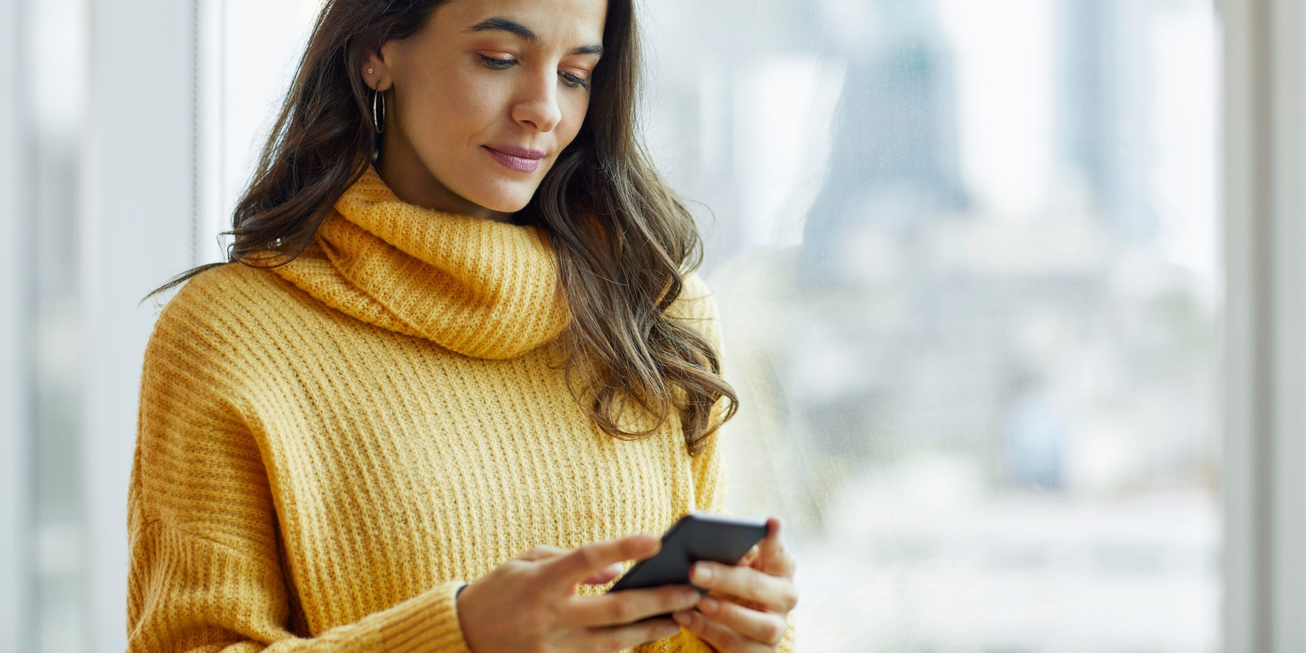 A woman in a yellow sweater uses a smart phone next to an office window.