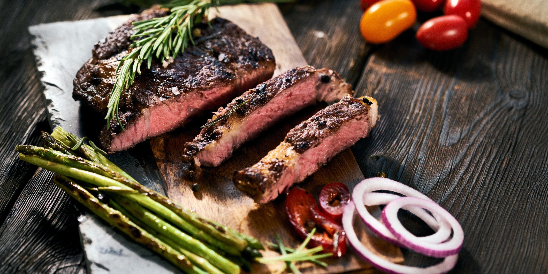 Sliced steak on a wooden cutting board topped with rosemary and accompanied by asparagus and sliced red onion