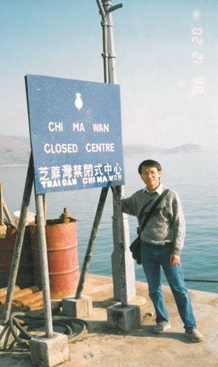 young man stands in front of a sign in vietnamese with water behind him