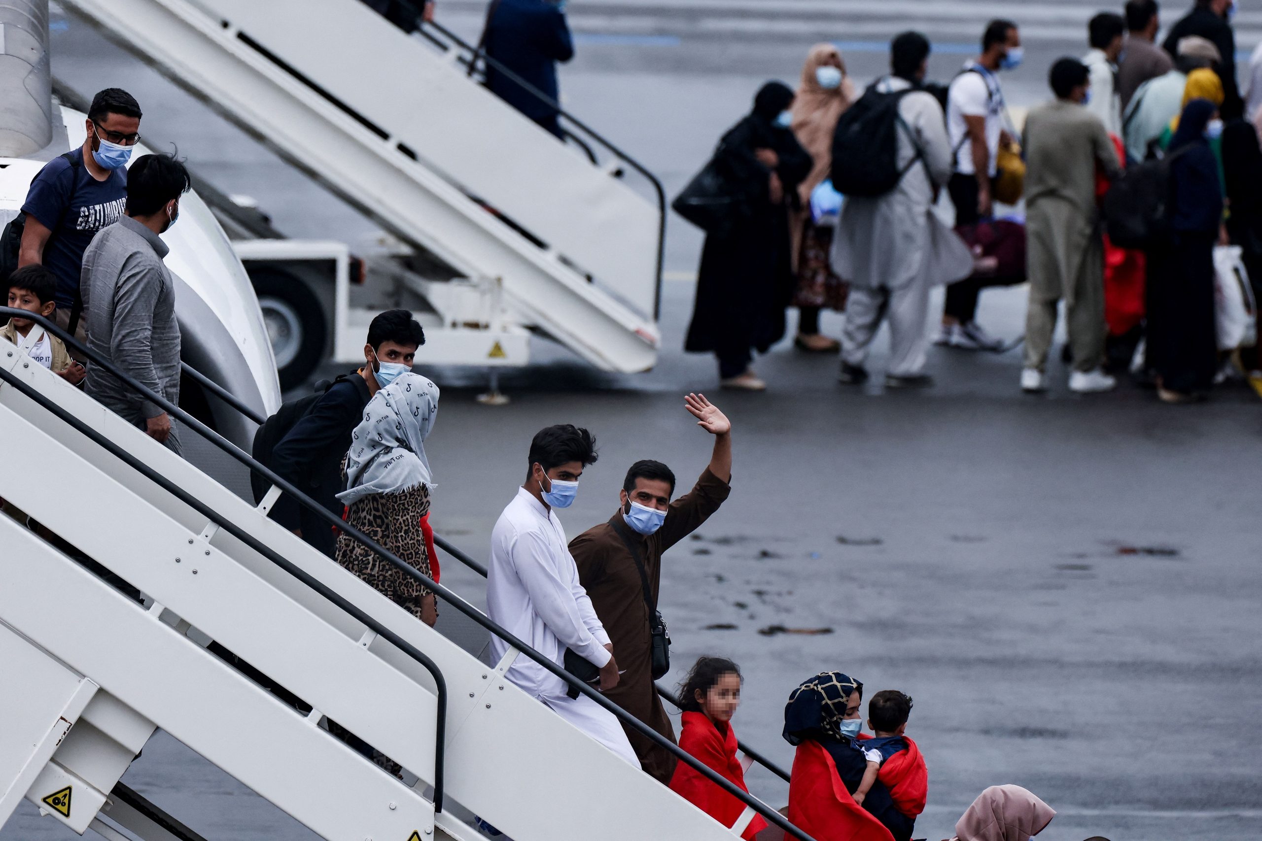 People descend the steps of an airplane wearing face masks while one waves to the camera.