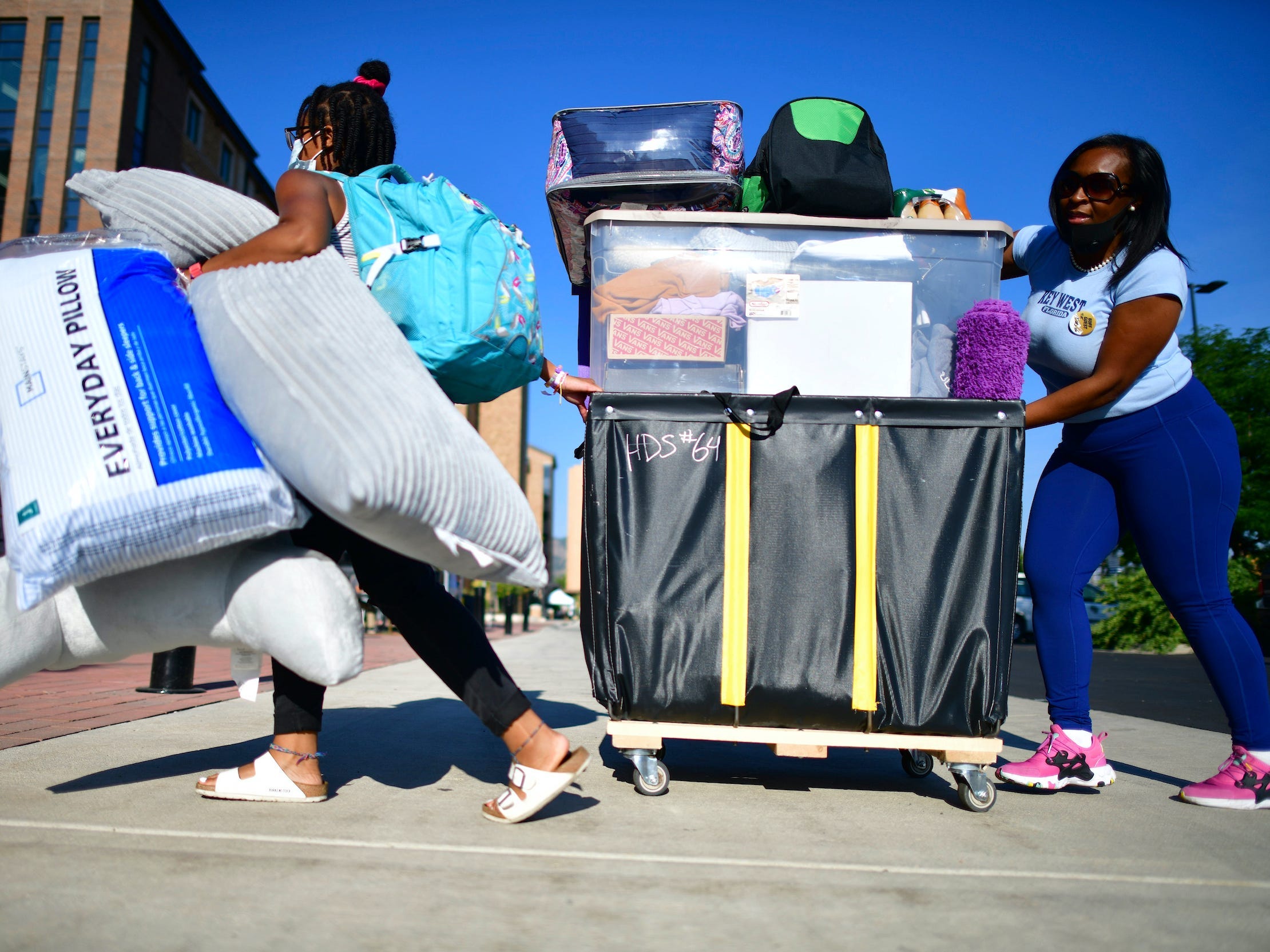 Woman carries pillows while another woman pushes cart behind her full of dorm supplies