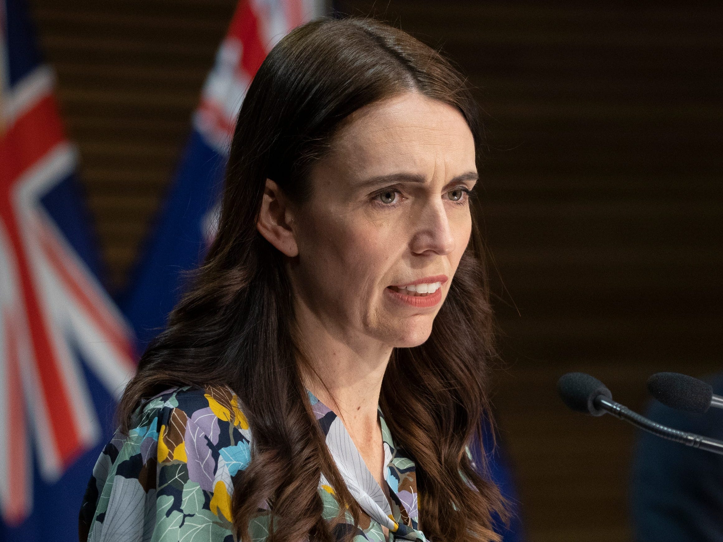 New Zealand Prime Minister Jacinda Ardern at a lectern with flag and a man in the background