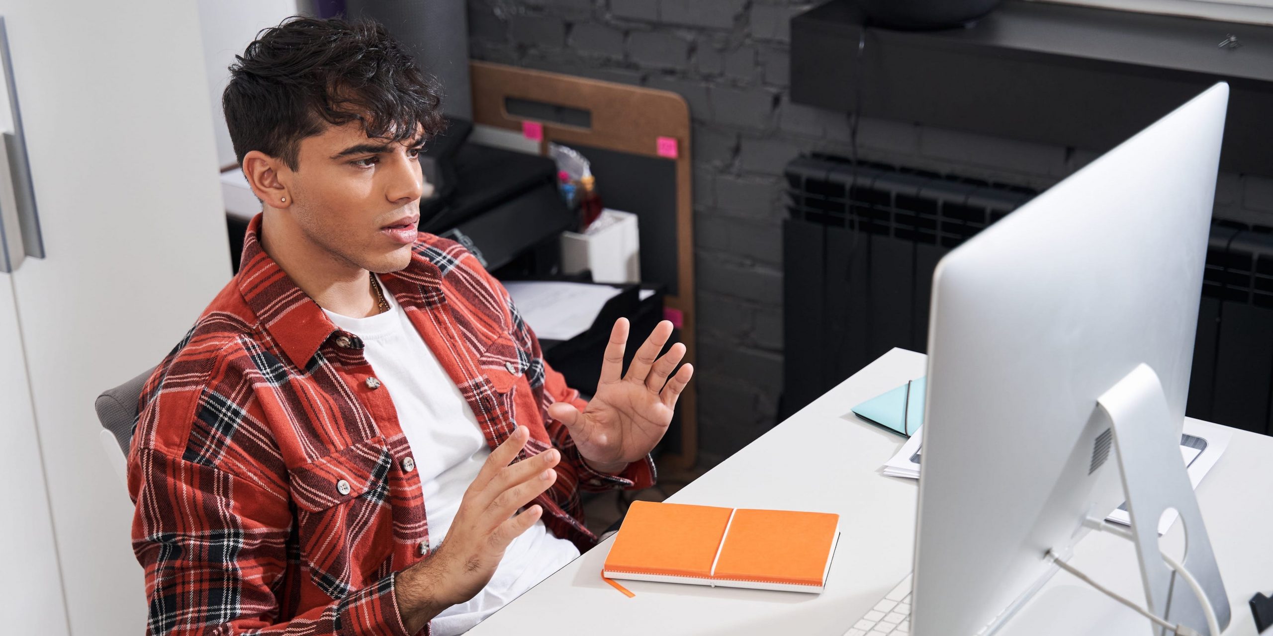 A high angle view of a man sitting at a desk in front of an iMac as he gestures.