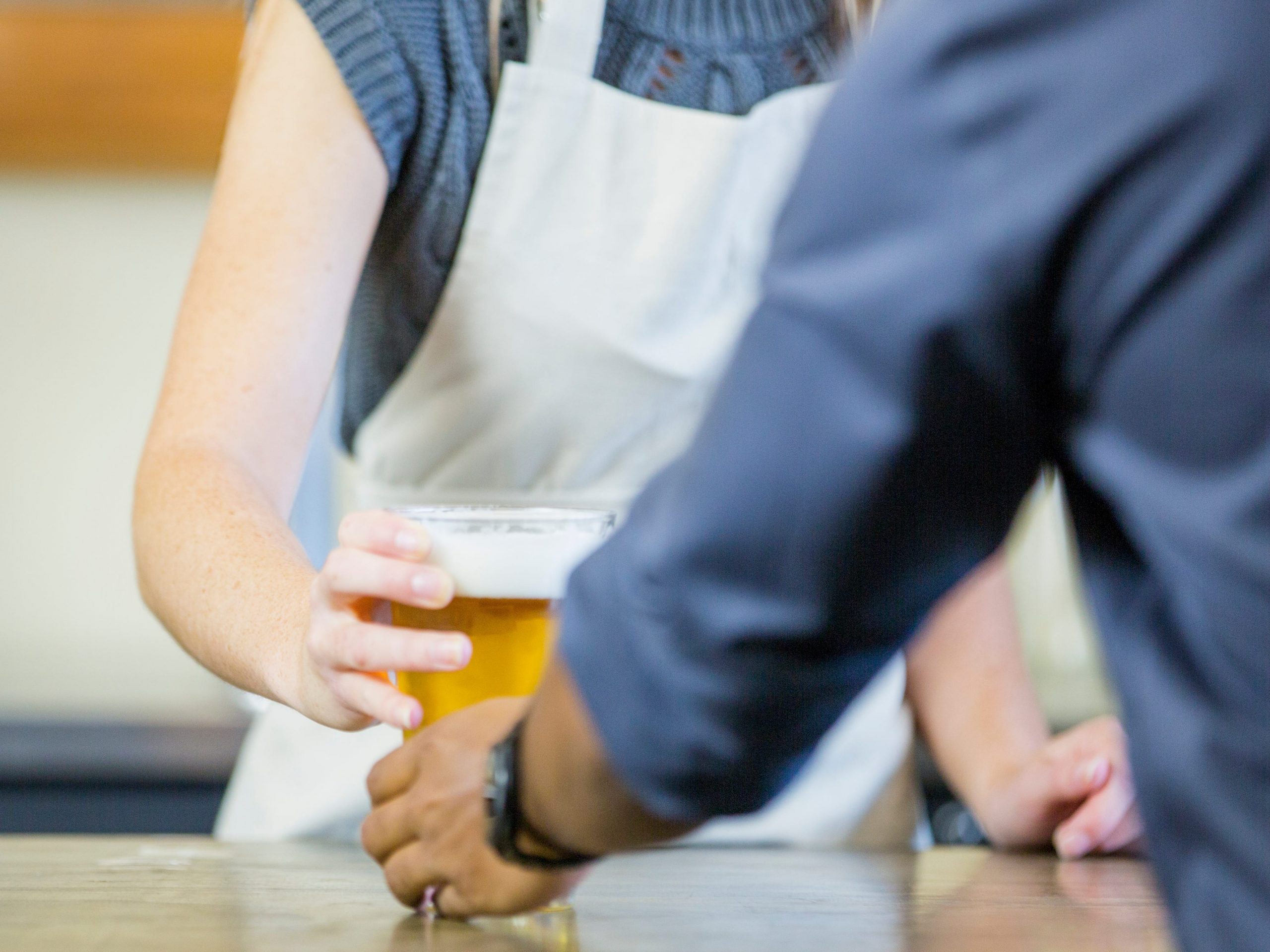 A female worker and man are indoors at a craft beer store. The woman is wearing an apron, and she is passing a glass of beer to the man.