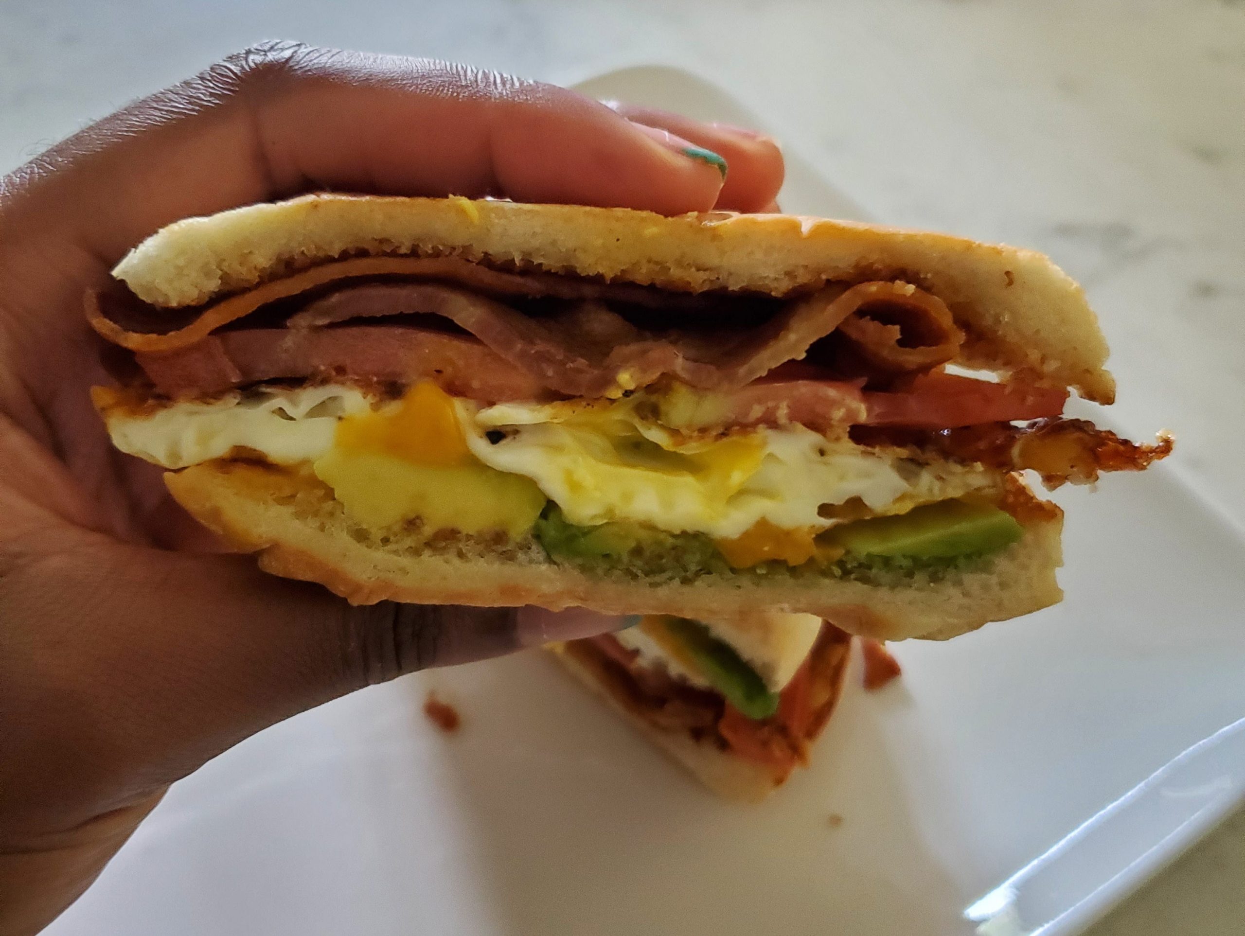 hand holding an egg breakfast sandwich with avocado and meat