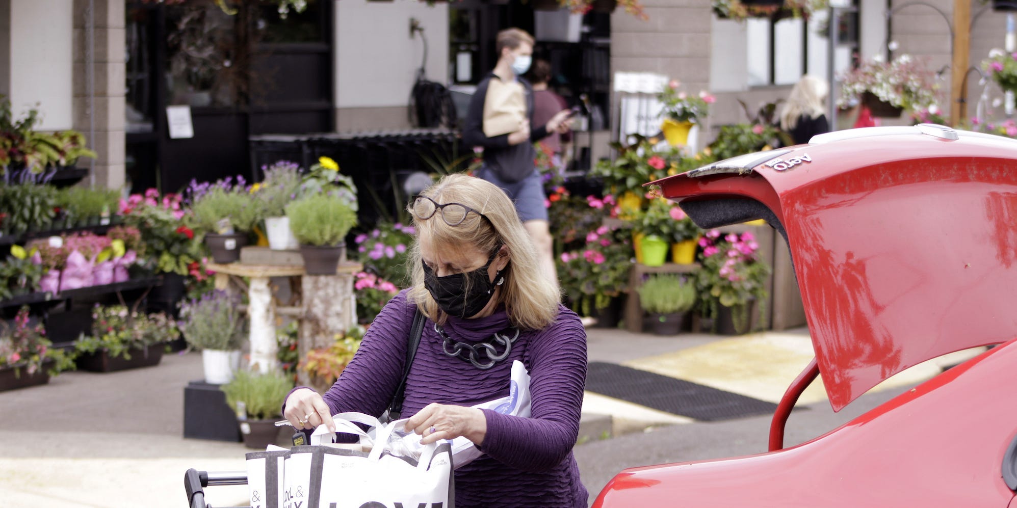 A woman wearing a purple top wearing a mask inspects her grocery shopping standing by an open trunk of a red car.