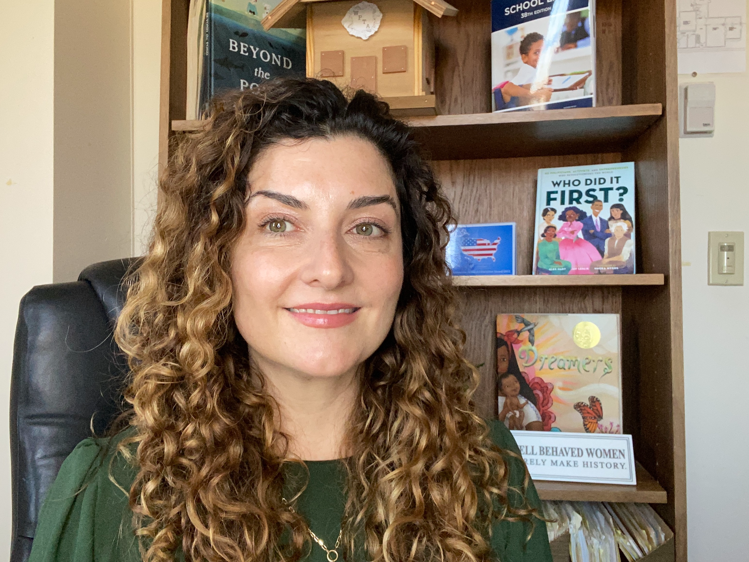 A woman with curly brown hair sits in front of a book case and smiles at the camera.