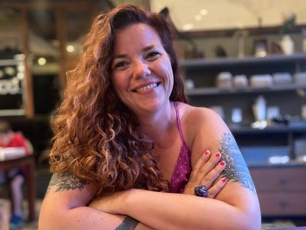 A woman with long red curly hair folds her arms and smiles at the camera.