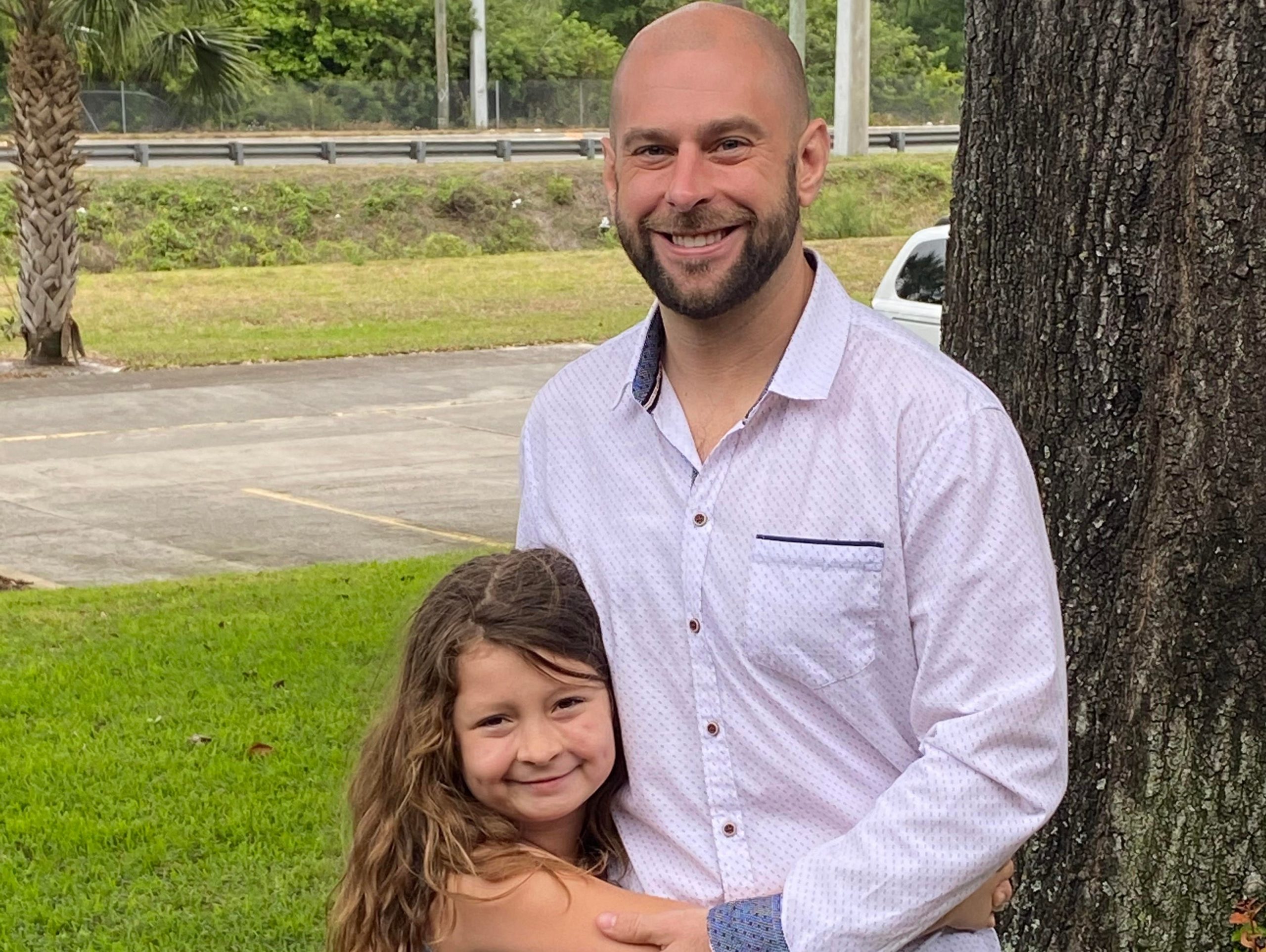 A man wearing a white shirt hugs his young daughter who wears a blue dress.