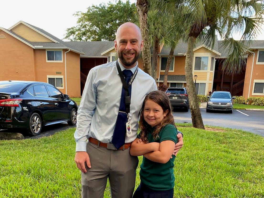 A man wearing a pale blue shirt and a dark tie puts his arm around his young daughter.