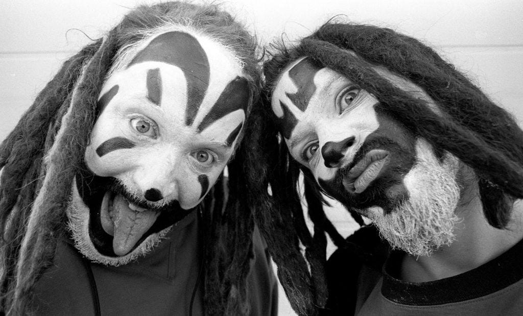 Two men with clown face paint and dark wigs make goofy faces at the camera in a black-and-white photo.