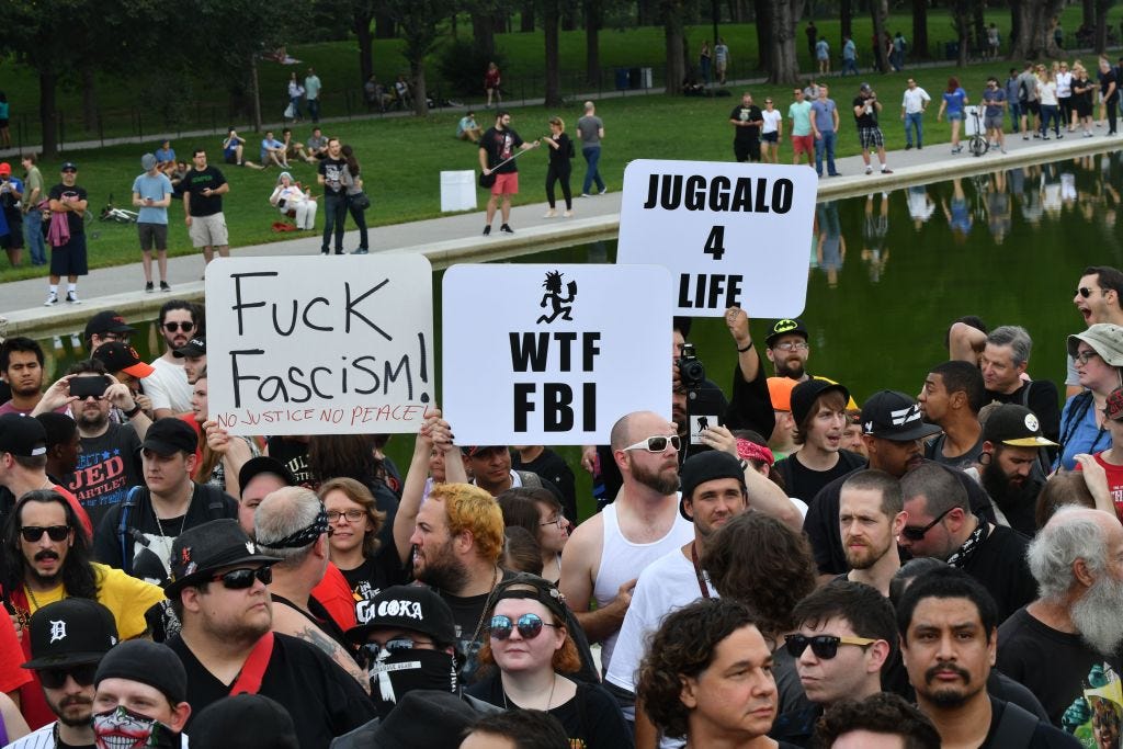 A group of Insane Clown Posse fans stands in a protest with signs that read 'JUGGALO 4 LIFE,' 'WTF FBI,' and 'Fuck Fascism.'