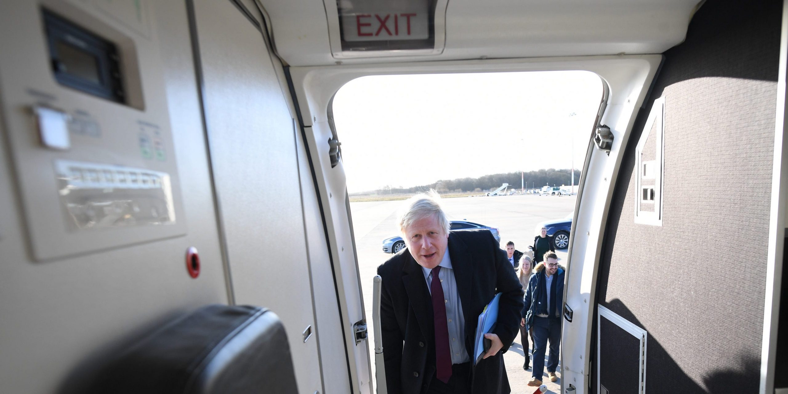 Boris Johnson gets on a plane. There is an exit sign above him.