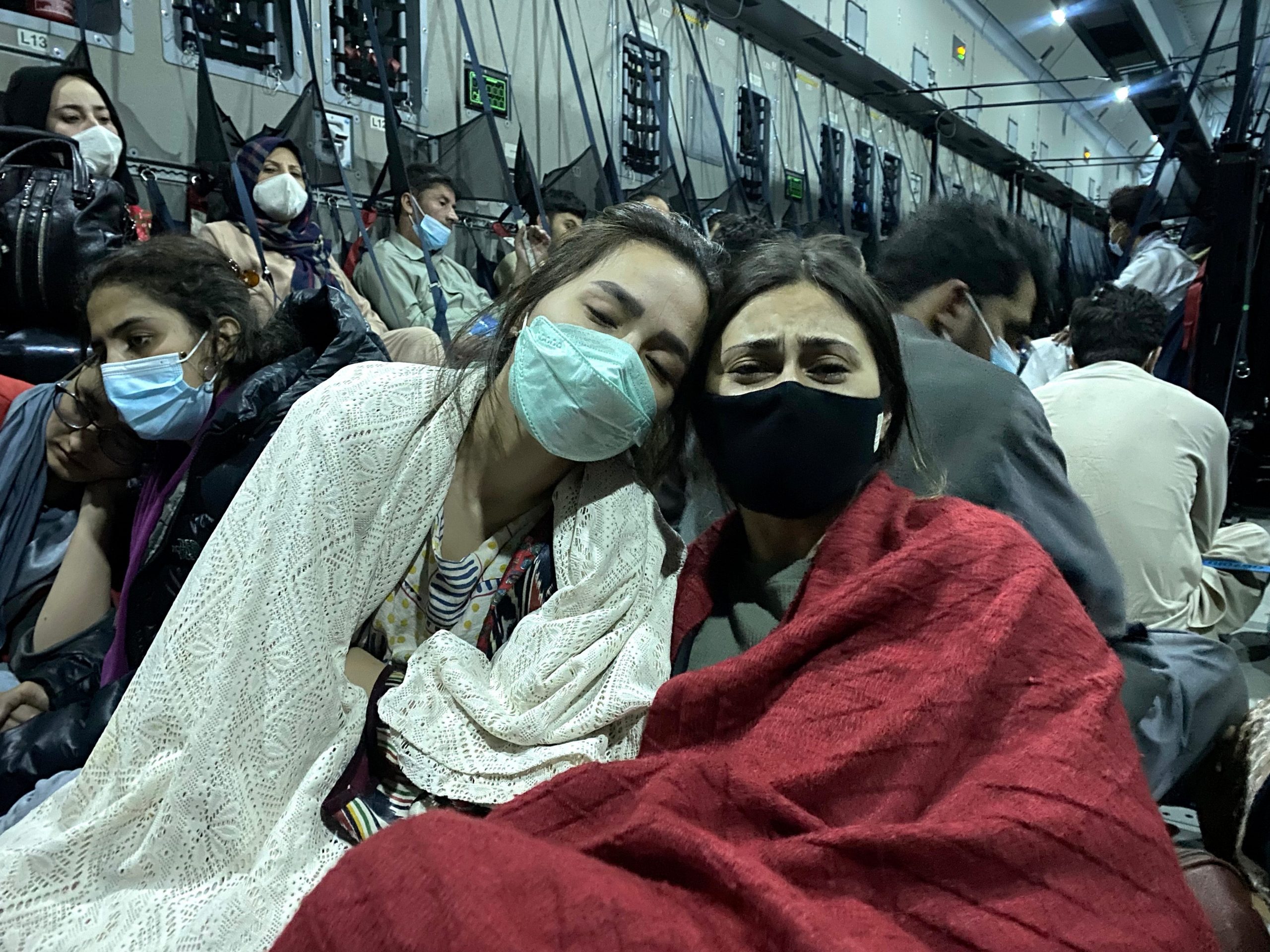 Two women, masked and draped in blankets, lean against one another amid a crowd of travelers.