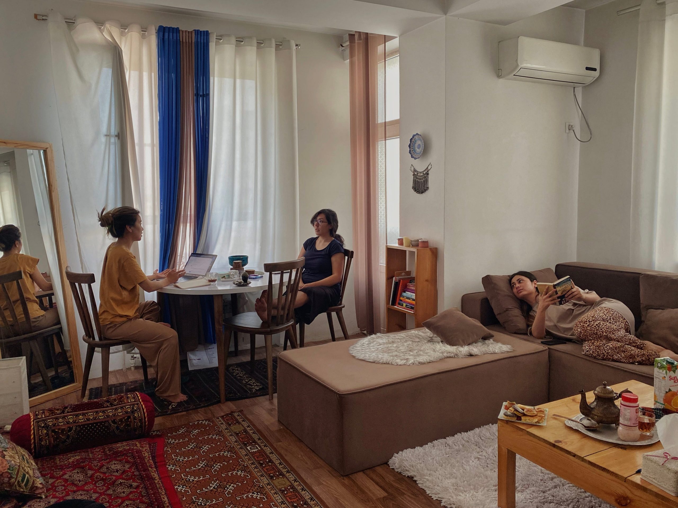 Two women sit at a table while a third lies on the couch.
