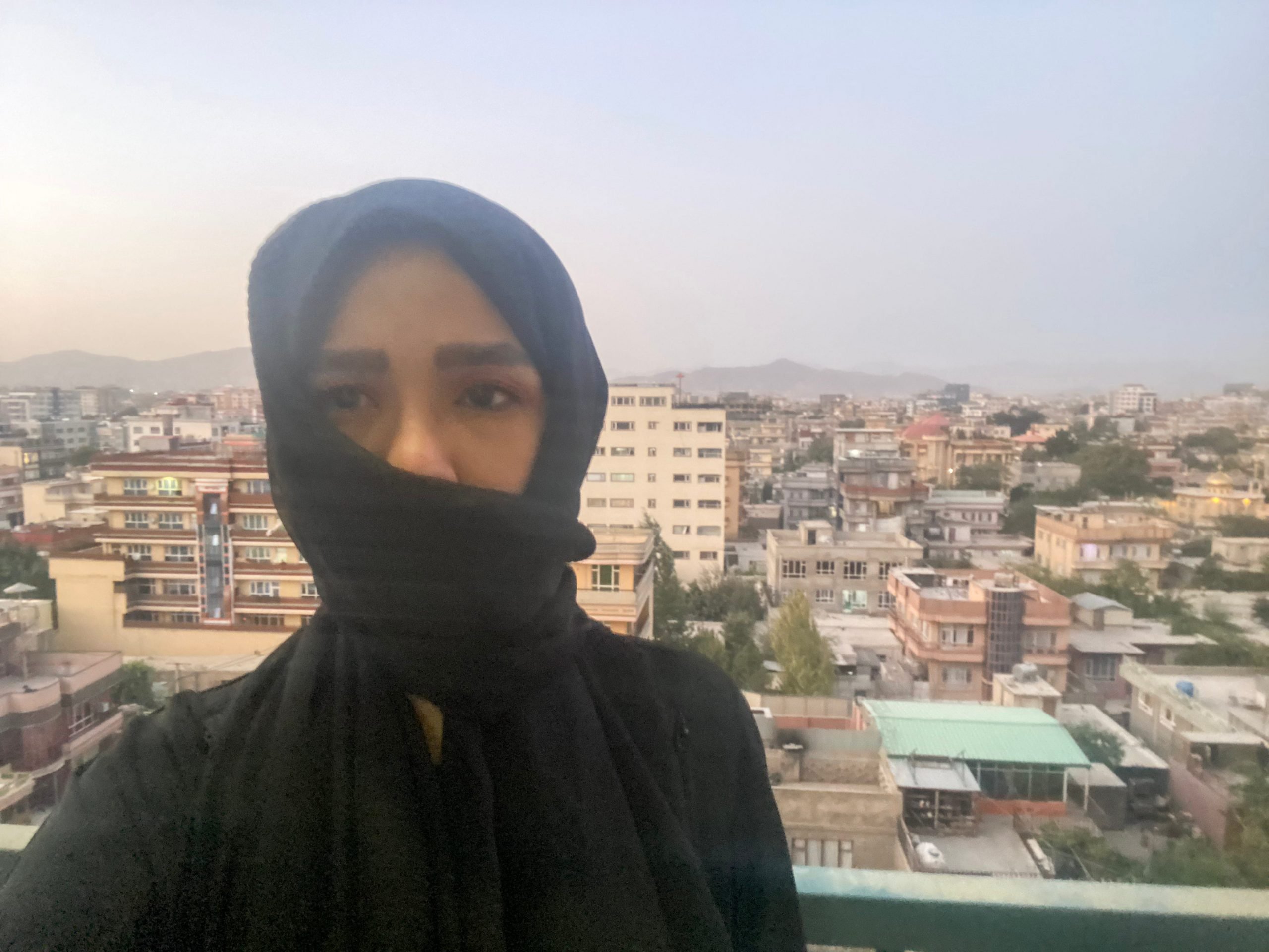 A woman, dressed in a black covering, poses in front of the Kabul skyline.