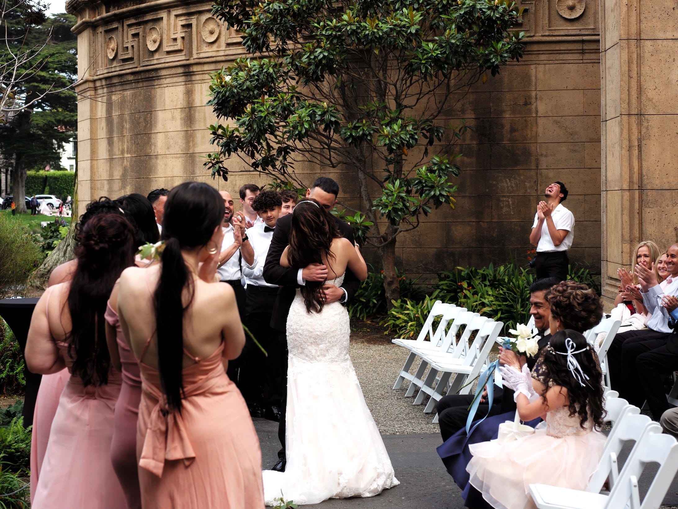 Bride and groom kiss at outdoor wedding ceremony while bridesmaids, groomsman clap